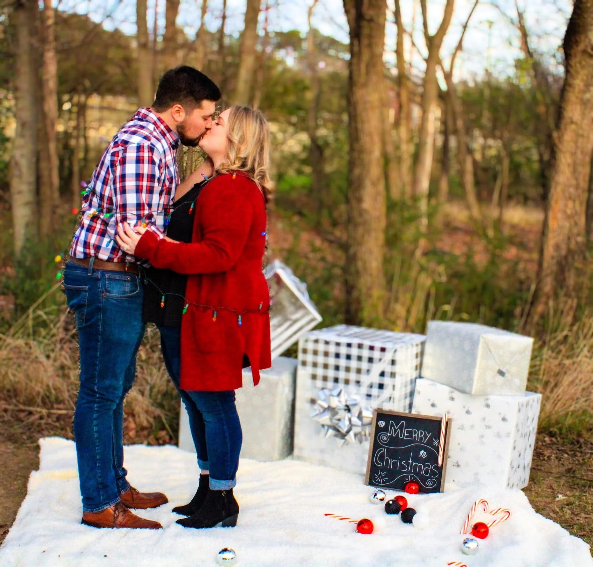 A couple kissing outdoors in a snowy forest near a decorated Christmas display with a chalkboard sign that says "Merry Christmas" and holiday ornaments.