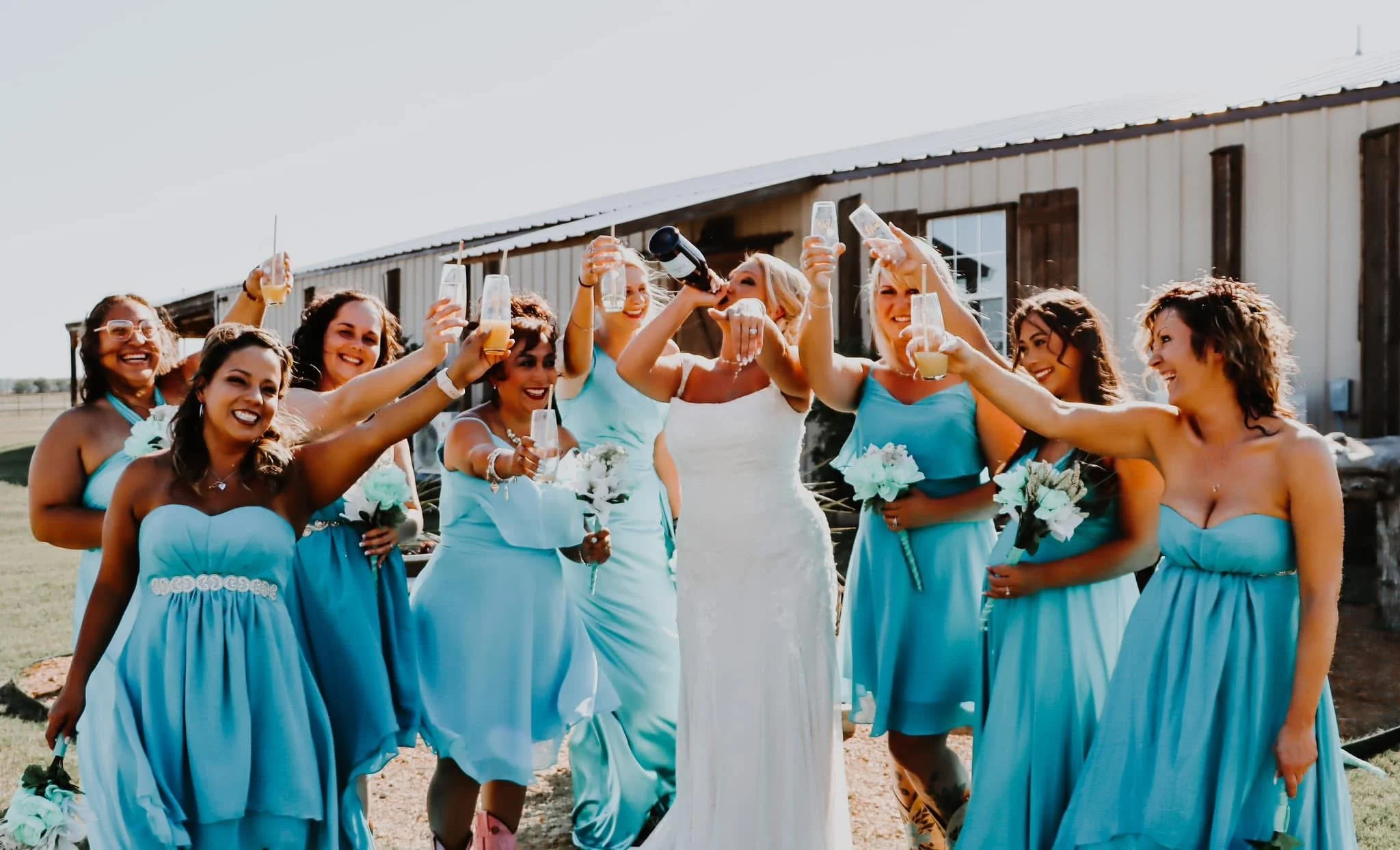 A group of women in blue bridesmaid dresses and a bride in a white wedding dress celebrating outdoors with drinks.