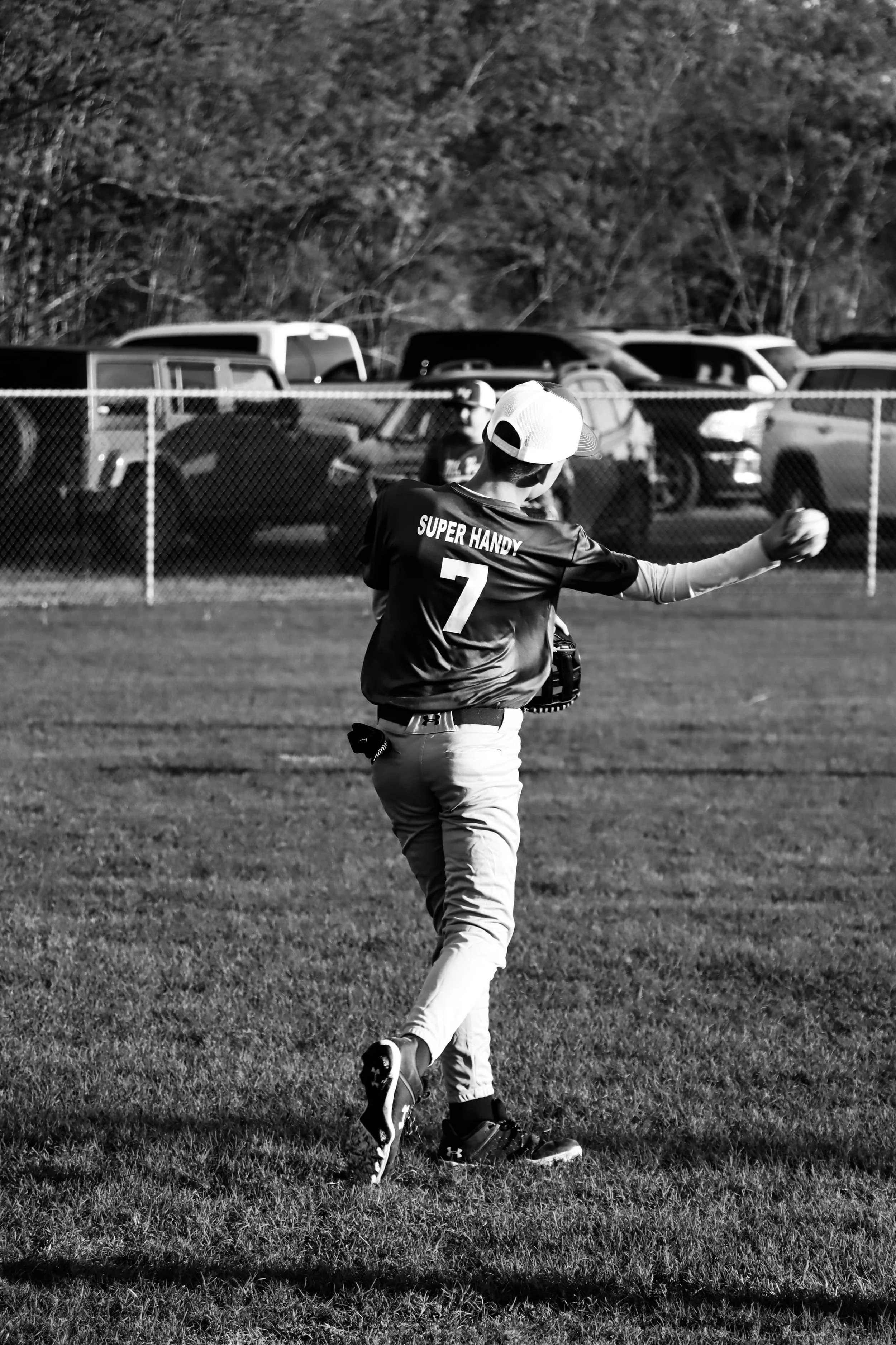A young baseball player wearing a jersey with the words 'Super Handy' and the number 7 on the back, practicing throws on a grassy field during the day. In the background, parked cars and a fence are visible, with trees behind them.