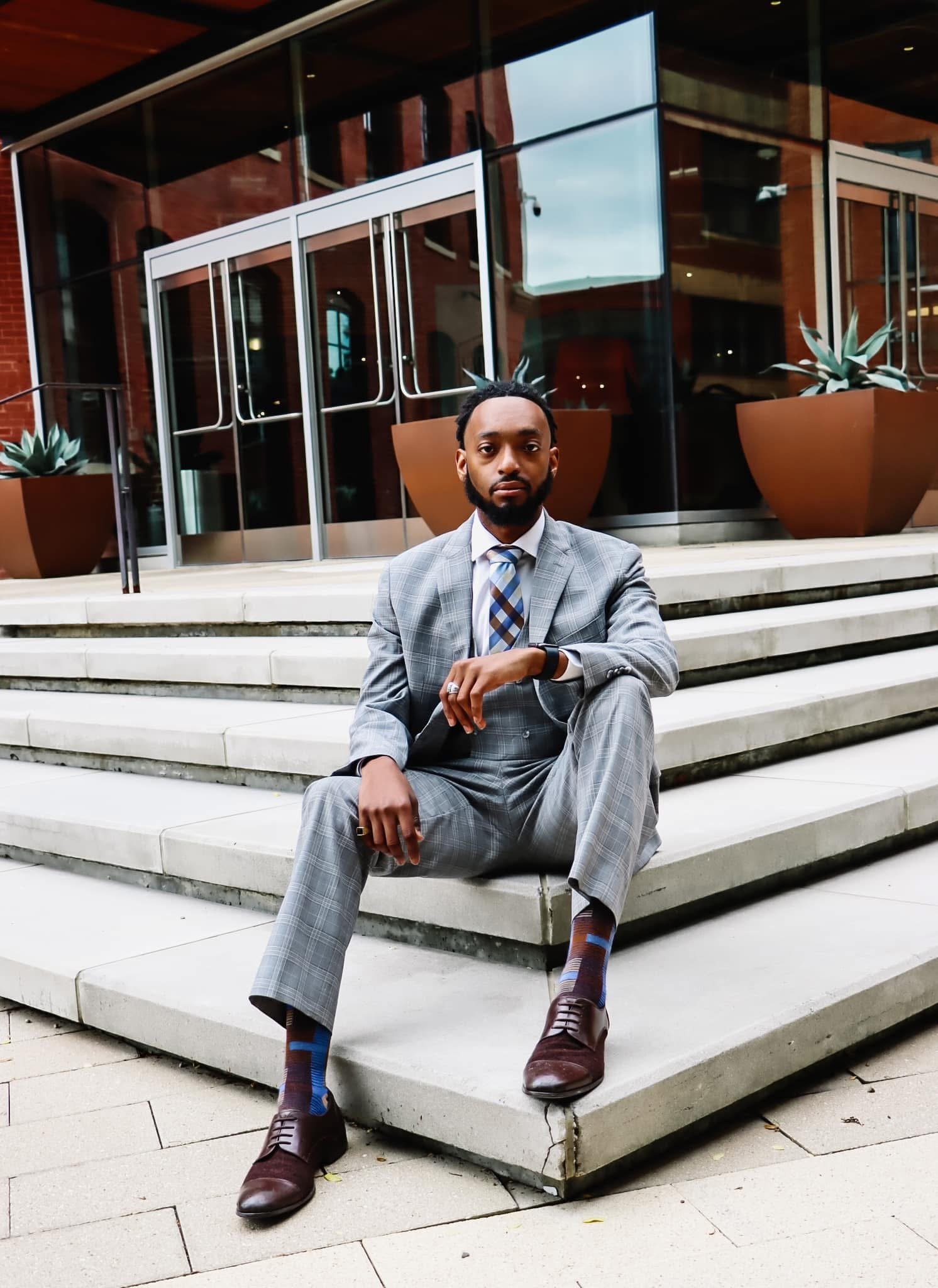 A man dressed in a gray checkered suit sitting on white stone steps outside a modern building with large glass windows and potted plants.
