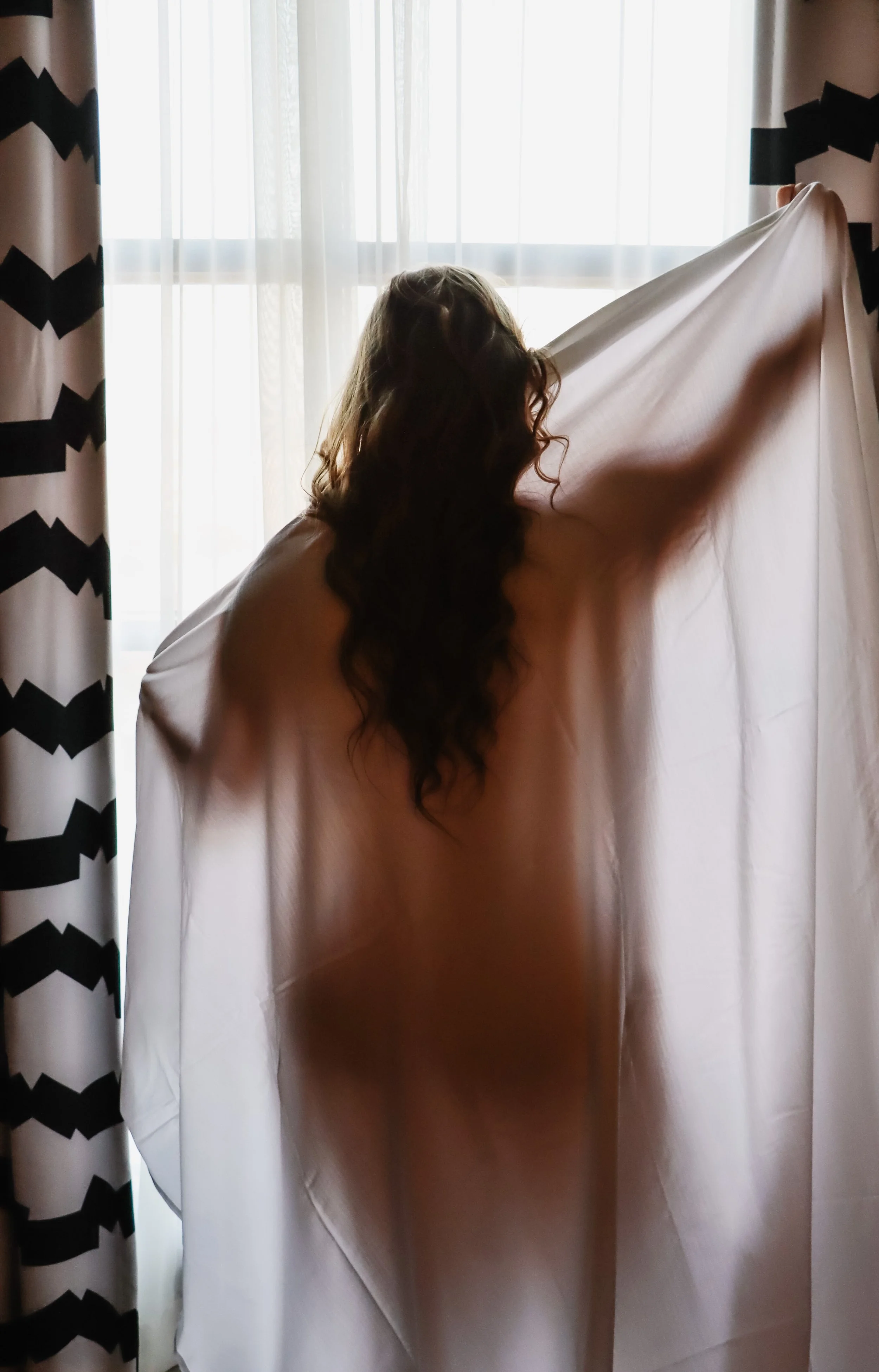 A person with long, curly brown hair holding up a light pink dress or fabric in front of a window with sheer curtains. Part of a curtain with black and white zigzag pattern is visible on the left side.