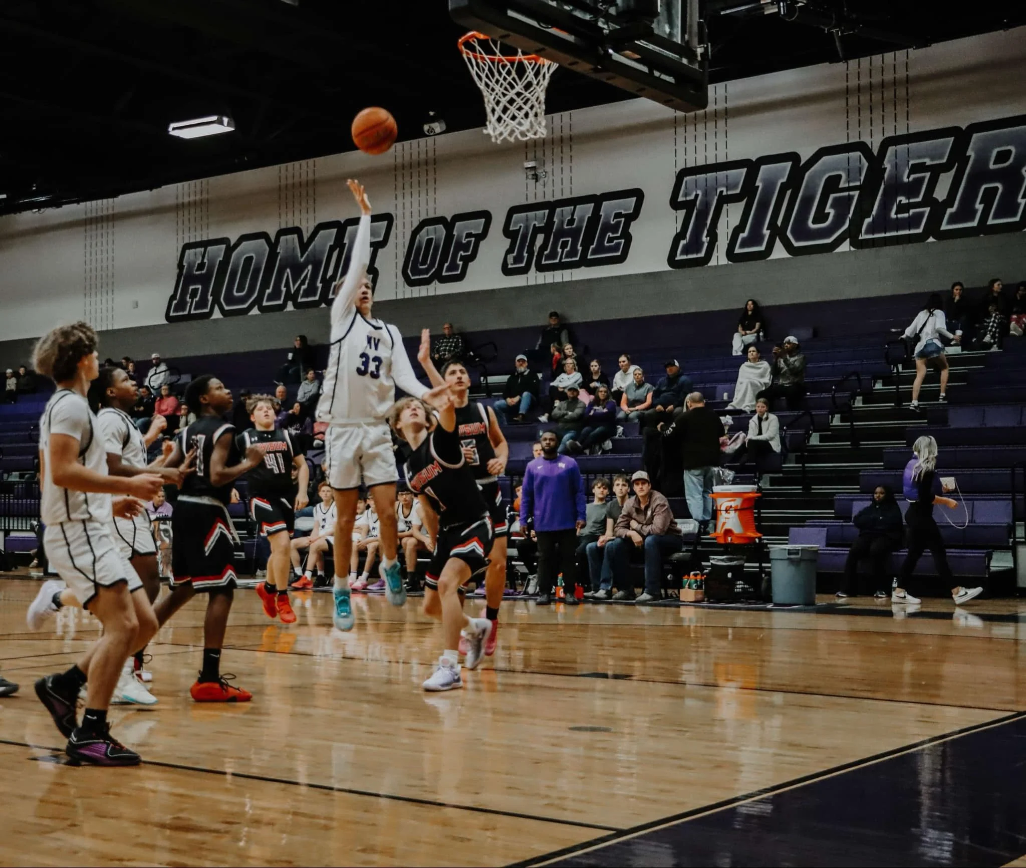 A basketball game in progress with players on the court and spectators in the stands, including a player shooting the ball towards the hoop.