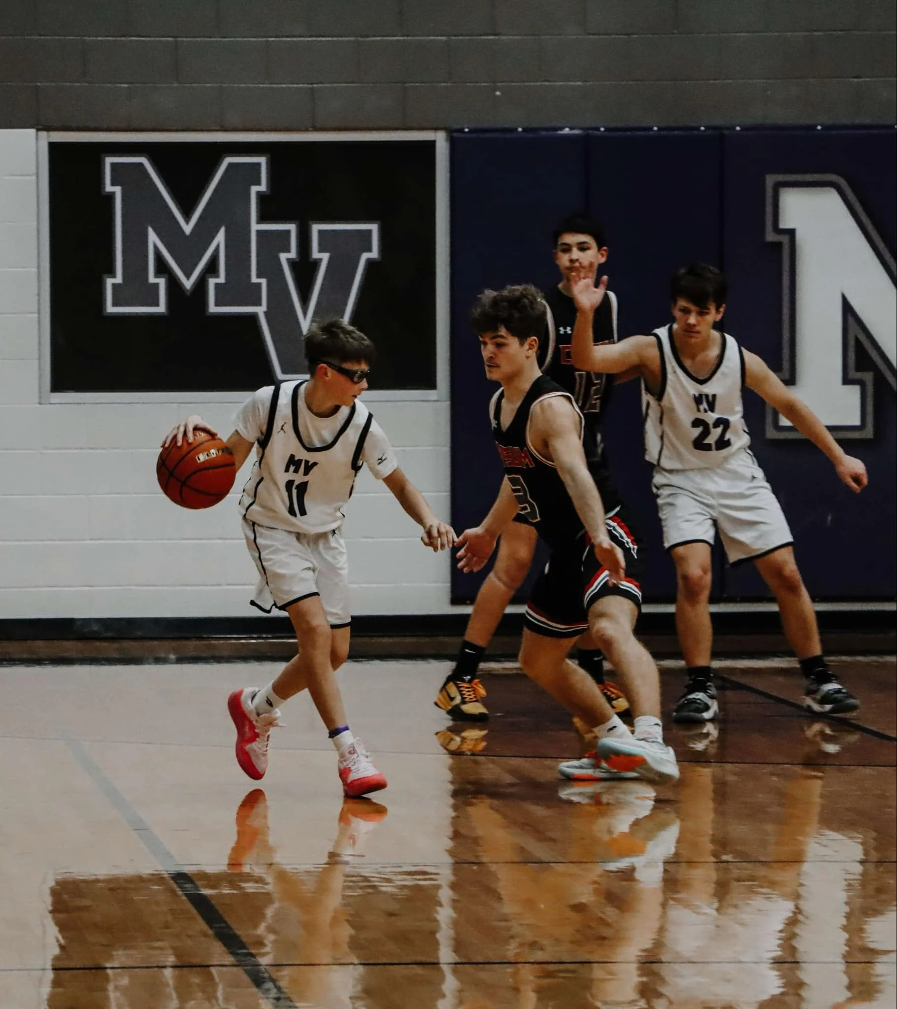 A group of young boys playing basketball on an indoor court, with some wearing white jerseys and one in black. One boy in a white jersey with the number 11 is dribbling the ball while others are positioned around him, including a boy in a white jerse