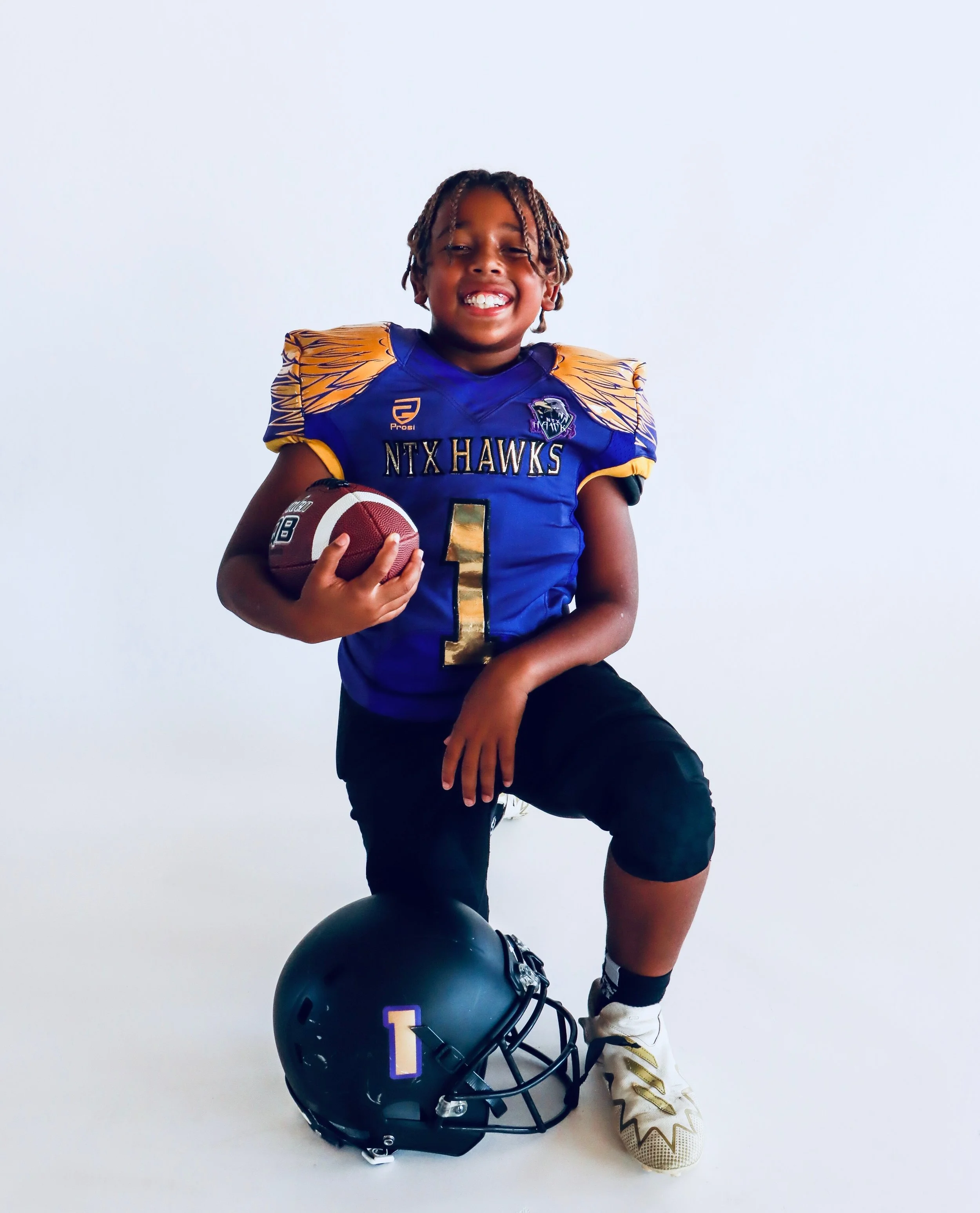Young boy in football uniform smiling, holding a football, with a black football helmet on the floor next to him, set against a plain white background.