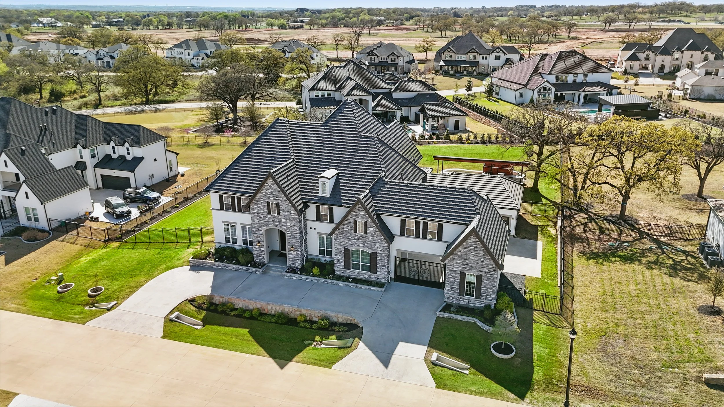Aerial view of a suburban neighborhood with large, modern houses, green lawns, trees, and a fenced back yard with a small swimming pool.