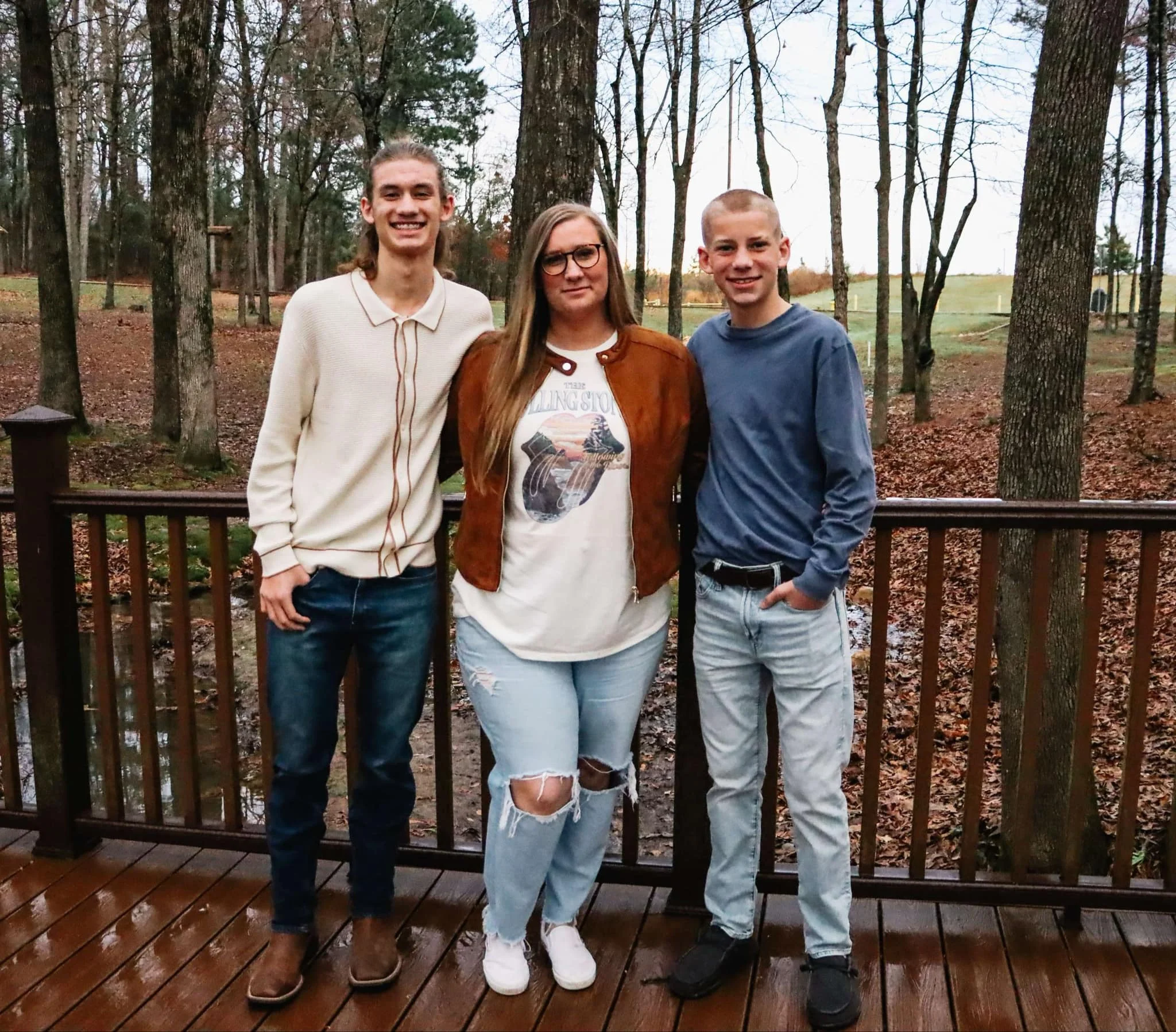 Three teenagers standing on a wooden deck outdoors surrounded by trees, smiling at the camera.