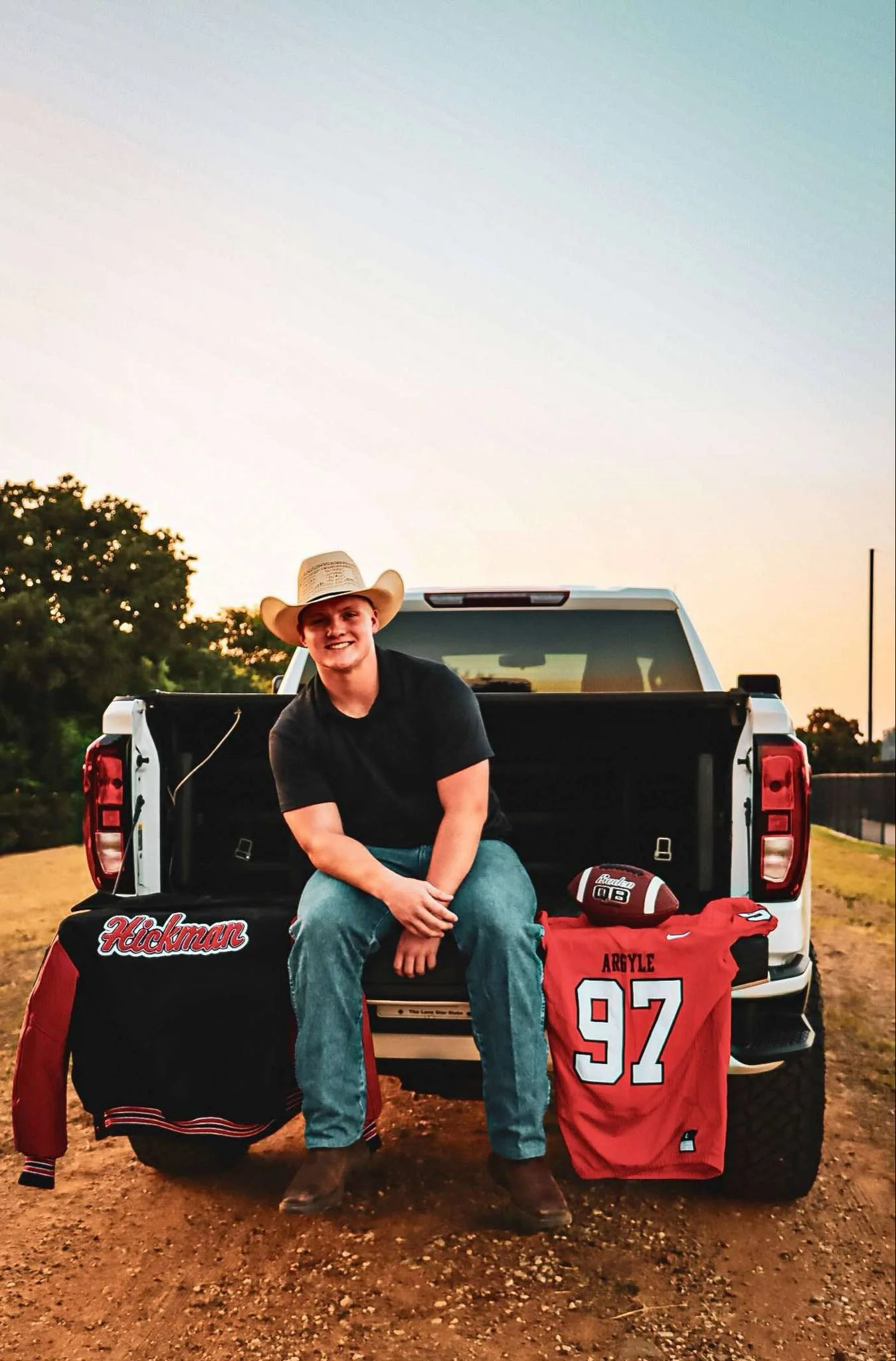 Young man wearing a cowboy hat sitting on the open tailgate of a white pickup truck during sunset. He is smiling and wearing a black T-shirt and jeans. Extra sports gear around him include a Hickman sports bag, a football, a red sports jersey with th