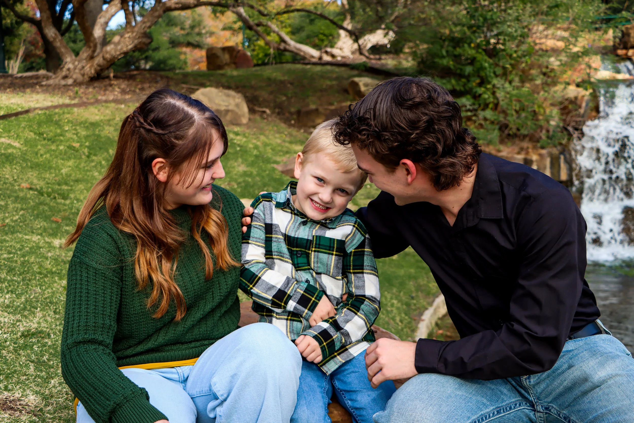 A family of four, consisting of a boy, a woman, and two men, sitting outdoors on grass near a small waterfall, smiling and looking at each other.