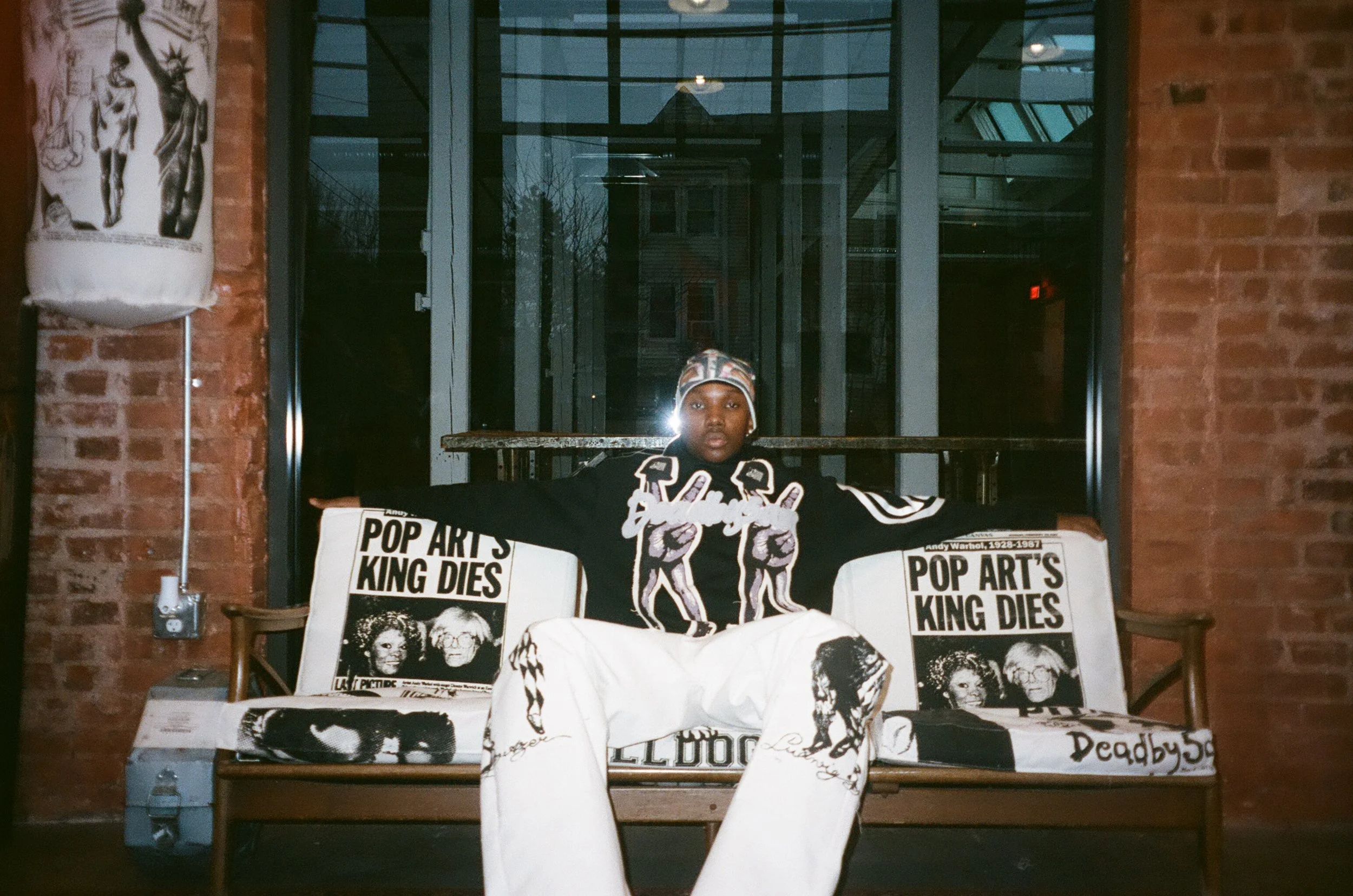 A person sitting behind a table covered with black and white posters featuring images of famous pop artists like Andy Warhol and The Beatles, in an industrial-style space with brick walls and large glass windows.