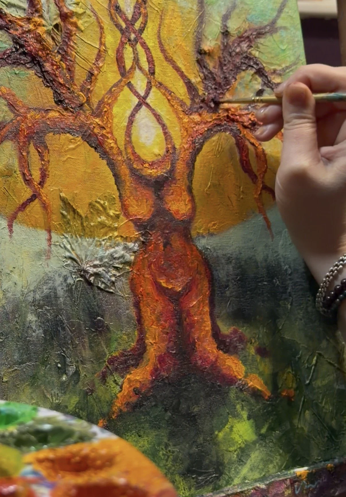 Close-up of an abstract painting of a fiery tree with orange, red, yellow, and black colors, textured with thick paint. Alicia Michelle is painting intricate lines on the canvas with a fine brush.