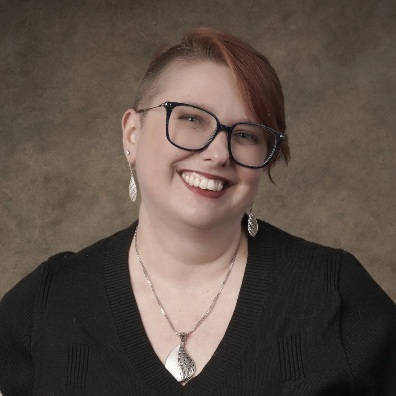 A woman smiling, wearing glasses, earrings, a necklace, and a black sweater, with short red hair and a brown background.
