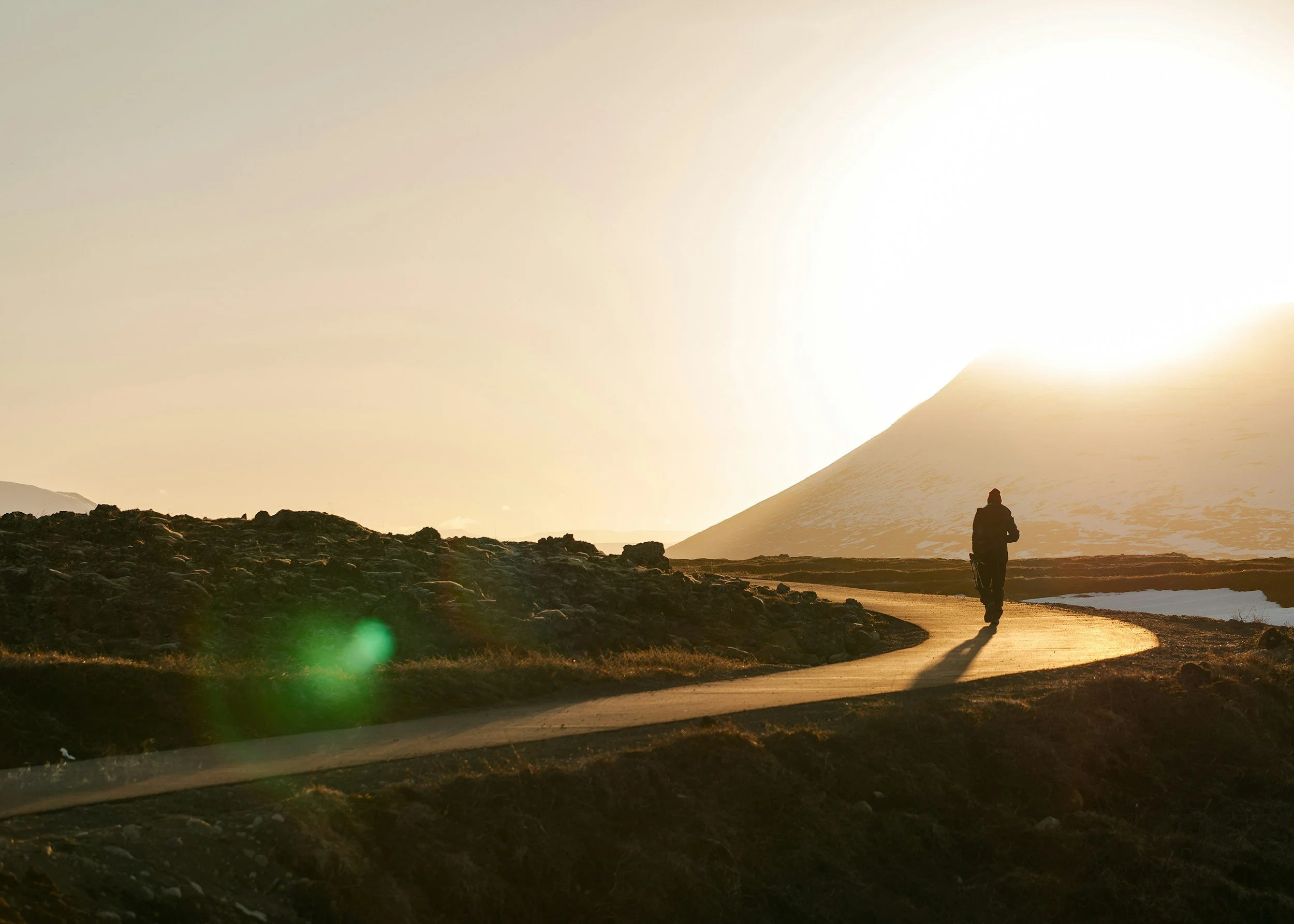 A person walking on a dirt path near a large snow-covered mountain with the sun setting or rising behind it, casting a glow over the landscape.