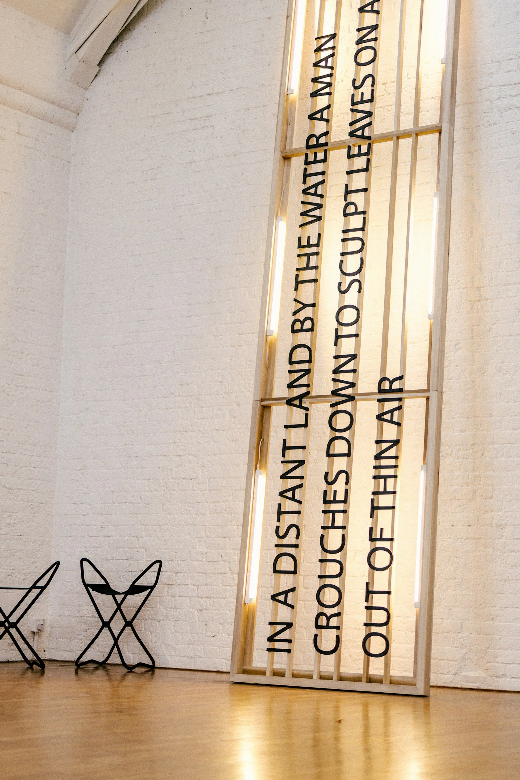 Interior view of a gallery or exhibit space with a white brick wall, black wireframe chairs, and a tall wooden frame with vertical black text on it.