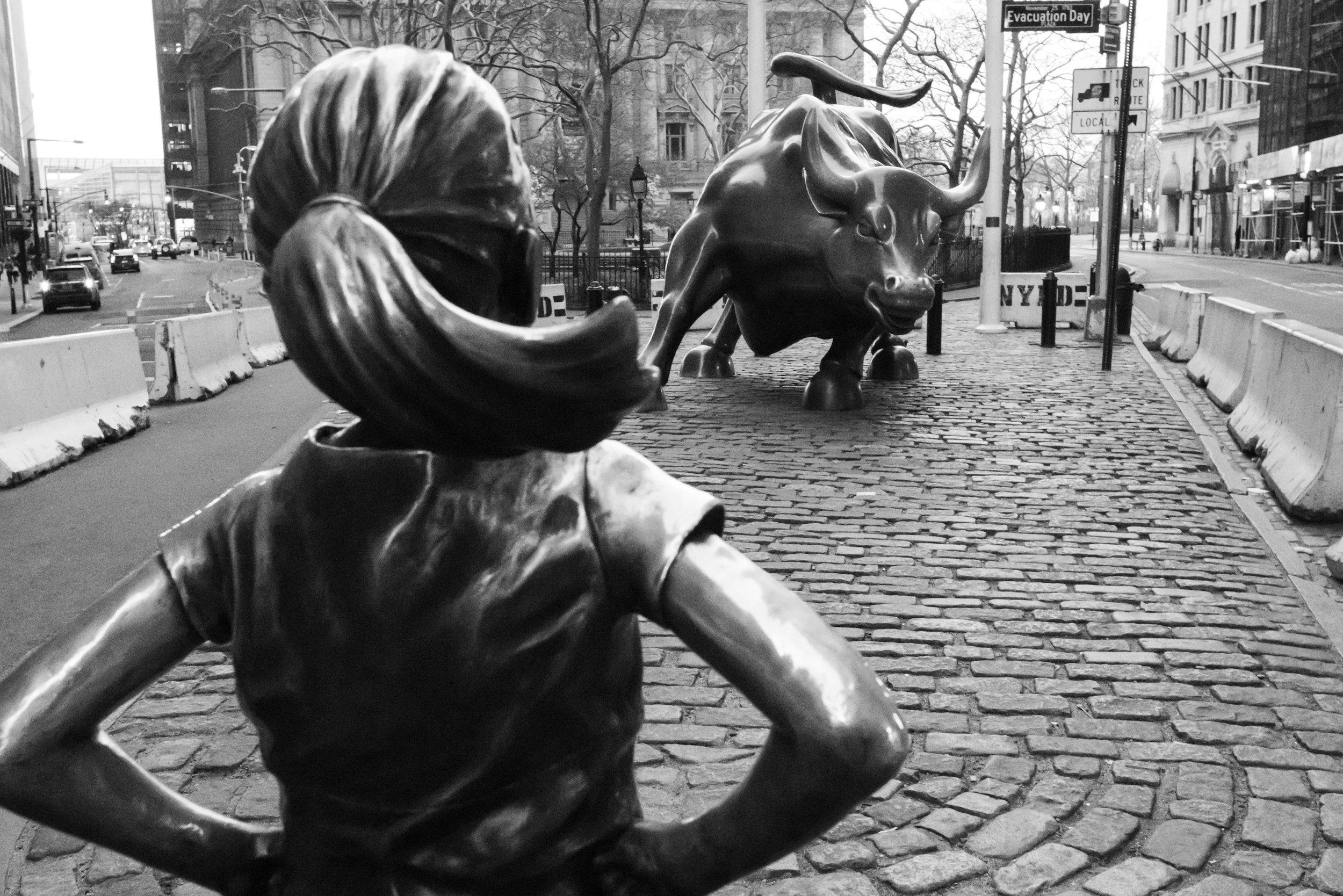 Black and white photo of the Charging Bull sculpture on Wall Street, with a child statue in the foreground, showing the back of the child with short hair and an arm extended, on a cobblestone street.
