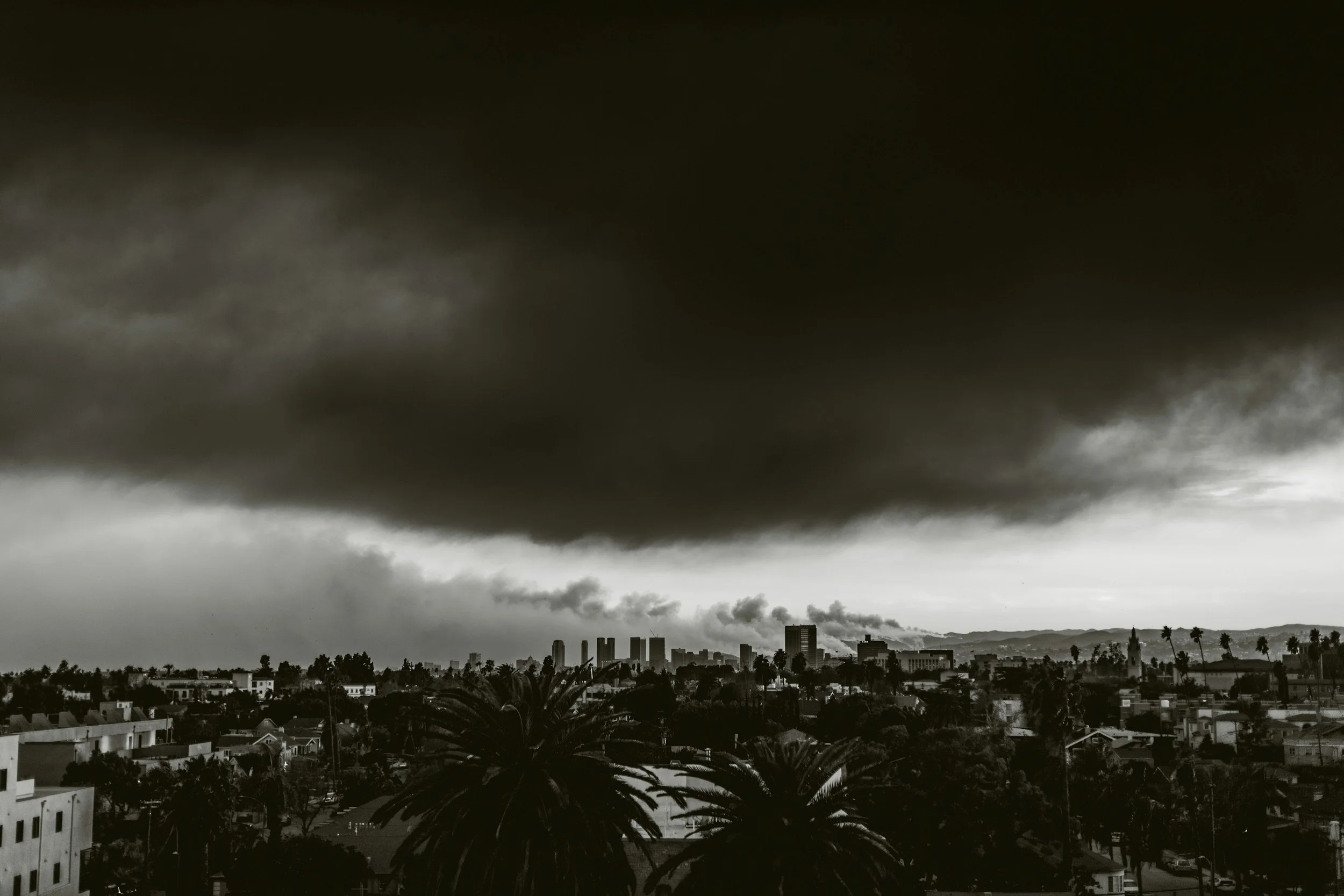 Dark storm clouds looming over a city skyline with smoke rising from industrial buildings and palm trees in the foreground.