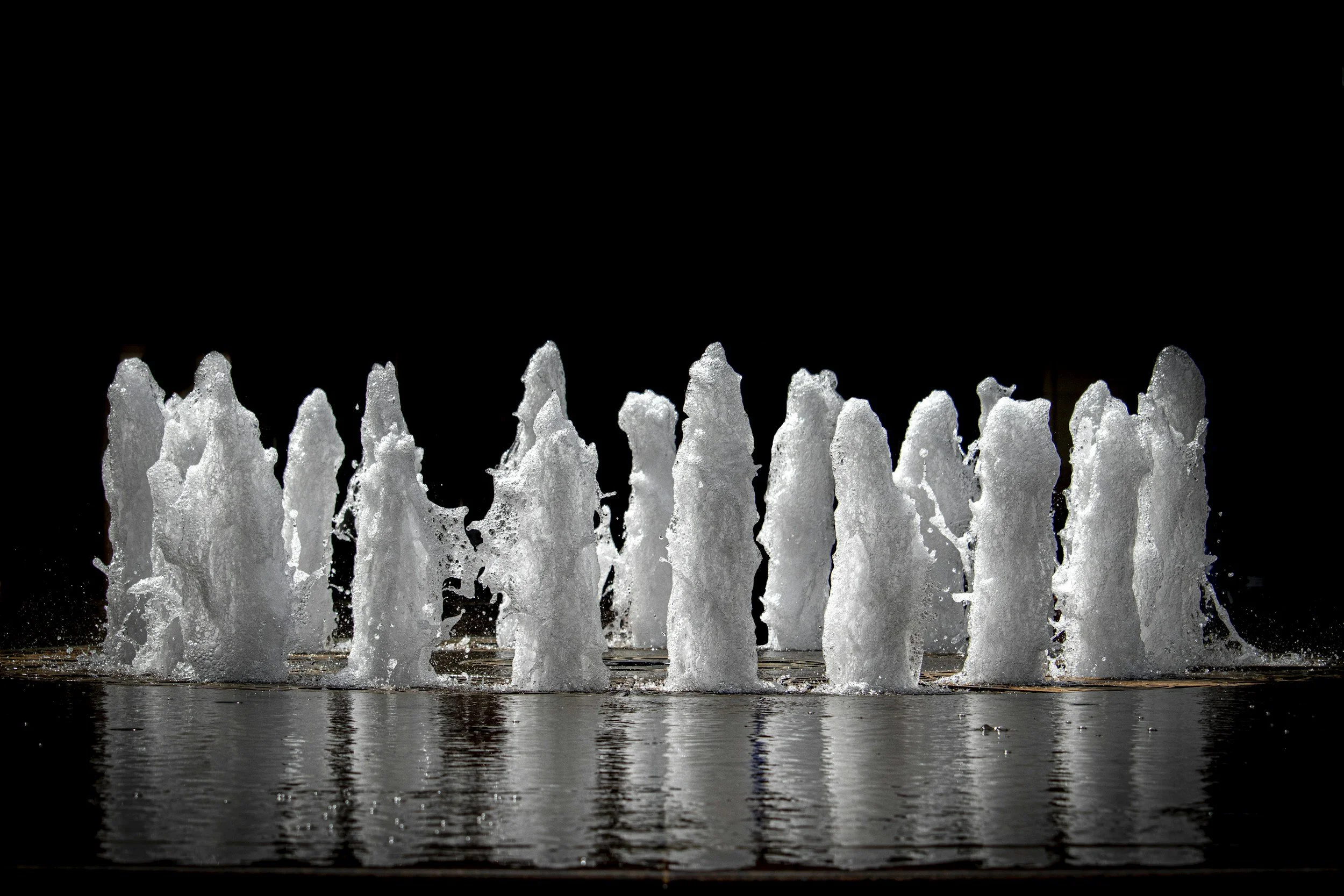A fountain with multiple water jets shooting upward, illuminated against a dark background, with water reflecting below.