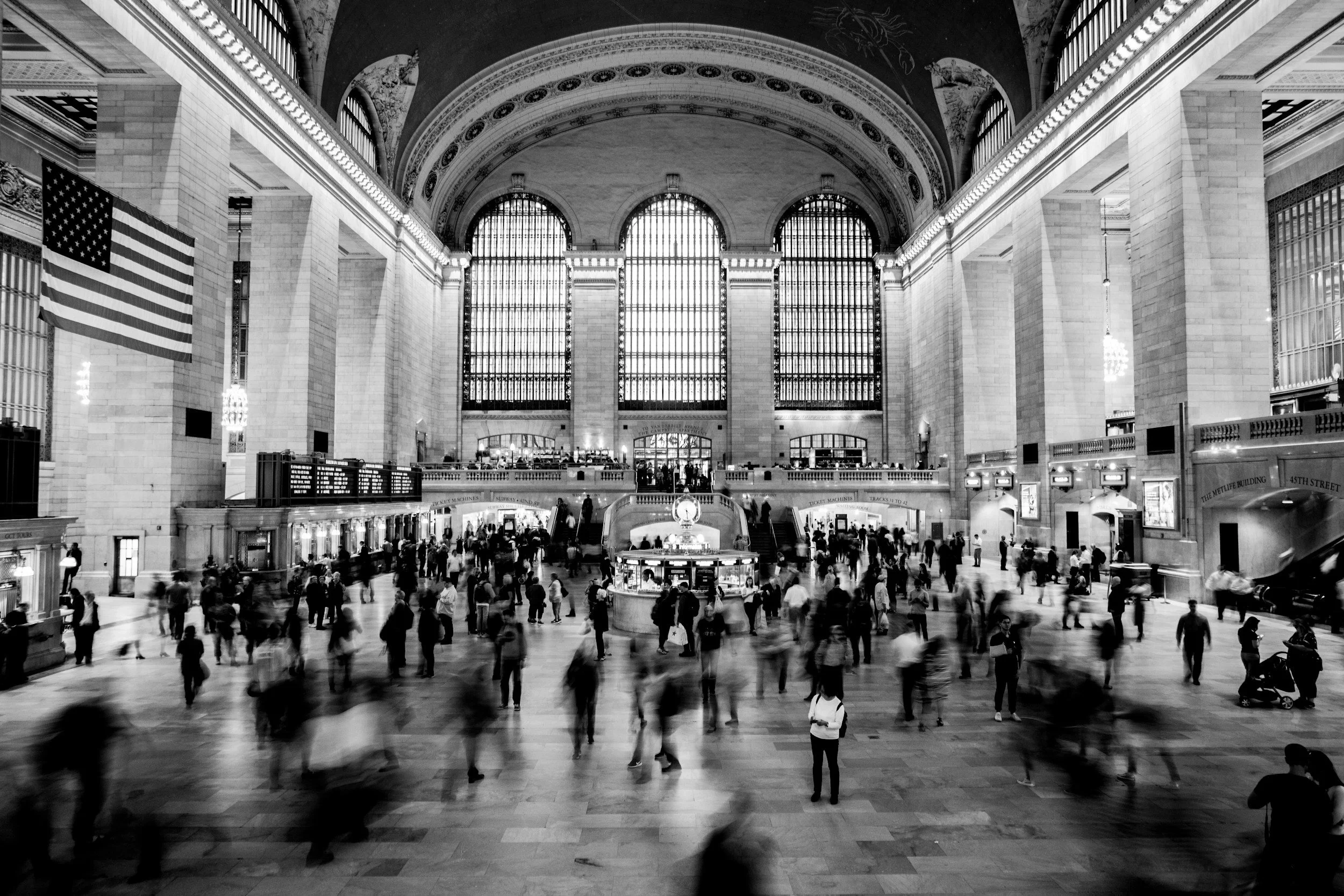 Black and white photo of Grand Central Terminal interior with large arched windows, high ornate ceilings, American flags, and busy crowd moving through the space.