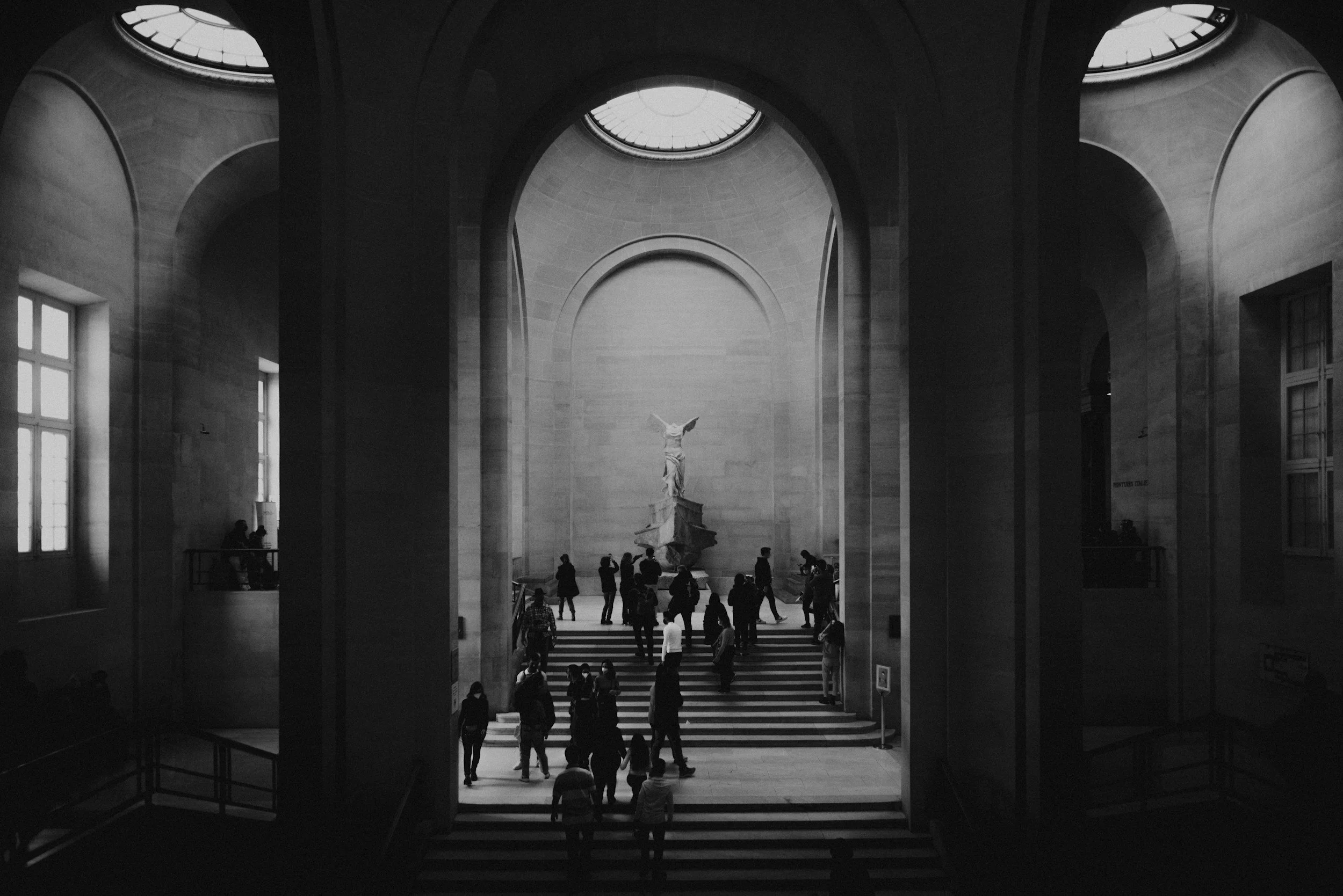 Interior view of a grand museum hall with high vaulted ceiling and large windows, featuring a statue on a pedestal and visitors walking around.