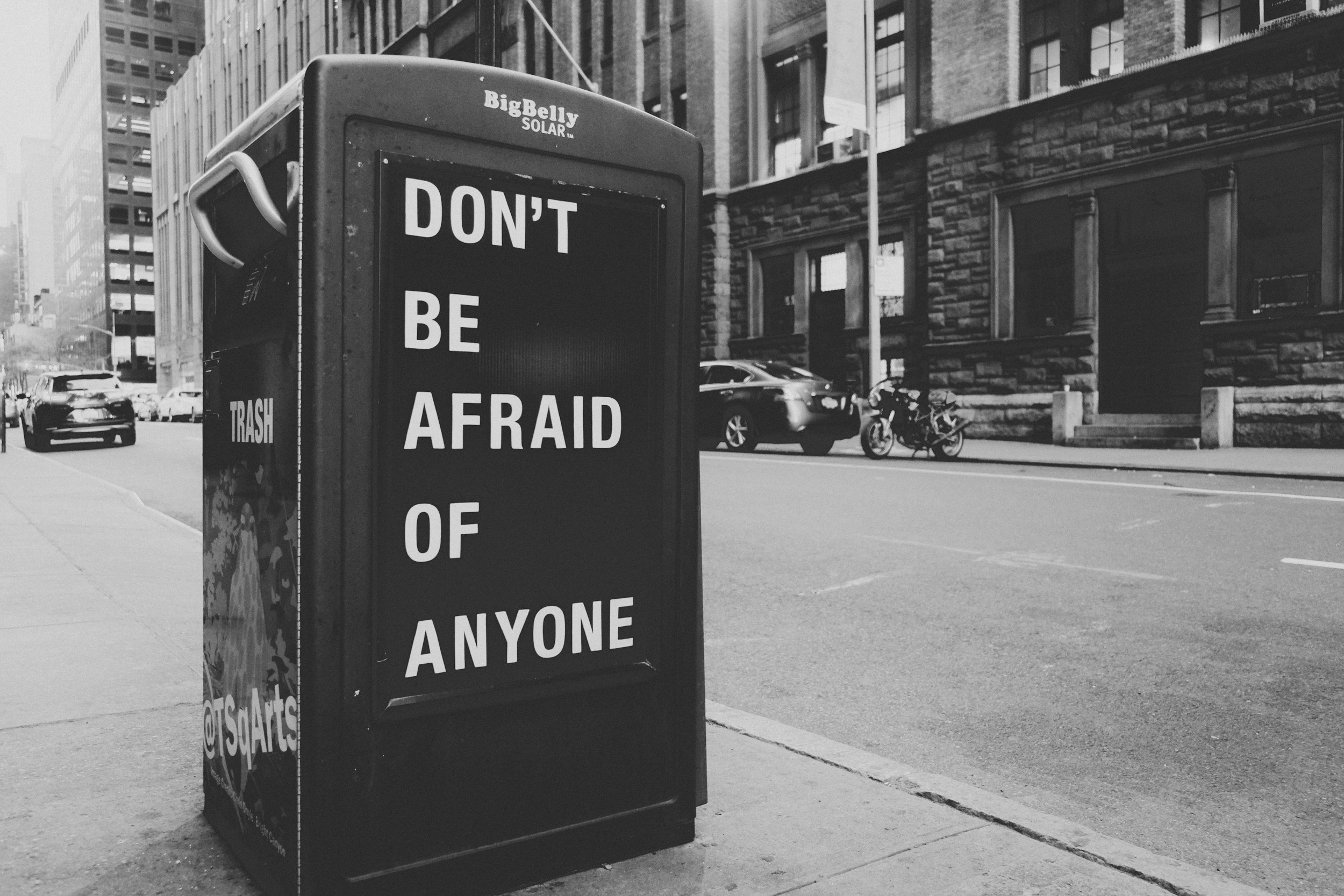A city street scene with a trash bin that has the message 'Don't Be Afraid of Anyone'. There are cars parked along the street and a building with brick and large windows in the background.