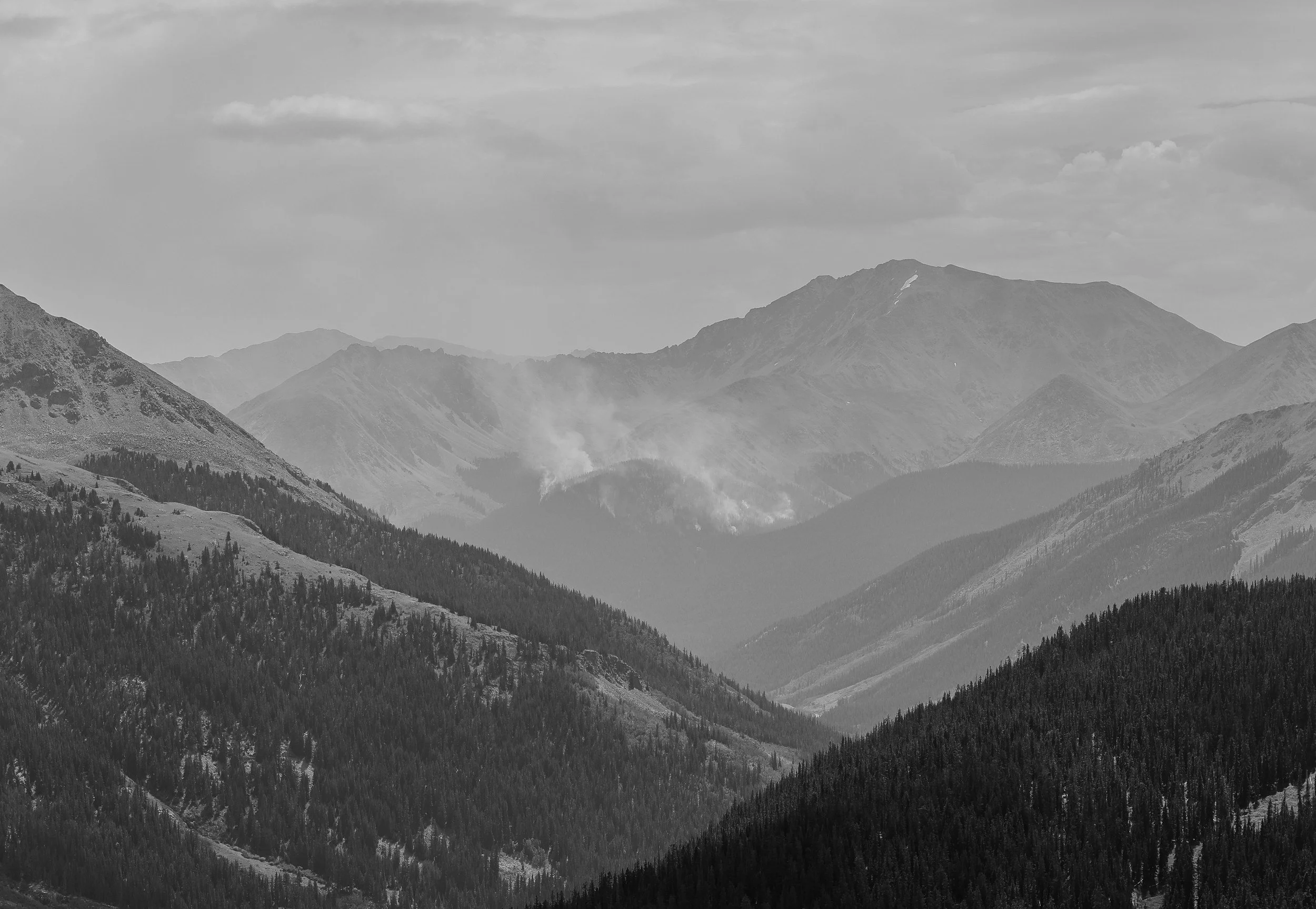 A black and white photo of a mountain range with multiple peaks and forested slopes, clouds in the sky.