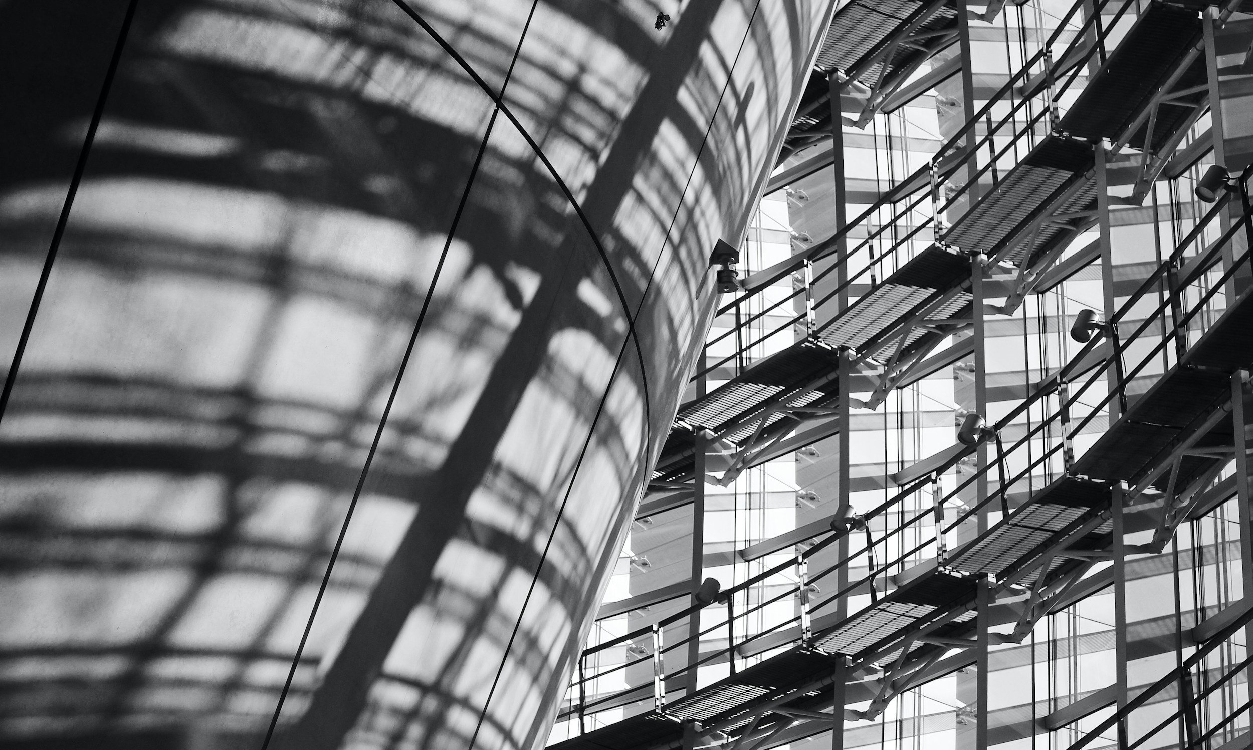 Black and white photograph of a modern building's interior, showcasing an abstract architectural design with shadows and patterns from metal stairs and glass windows.