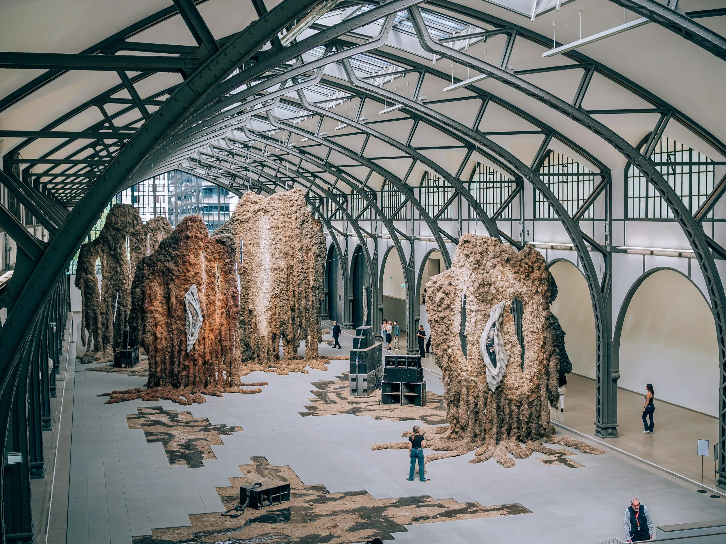Interior of a large building with a high arched ceiling and large windows, featuring several large, irregularly shaped rocks on the floor and a few people observing or walking around.