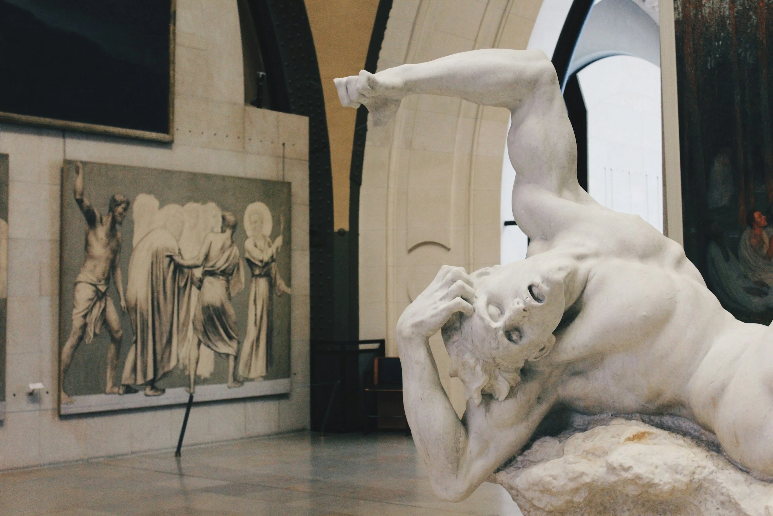 Close-up of a white marble sculpture of a woman lying on her side with her hand on her face, in a museum. In the background, there is a bas-relief depicting figures in classical attire.