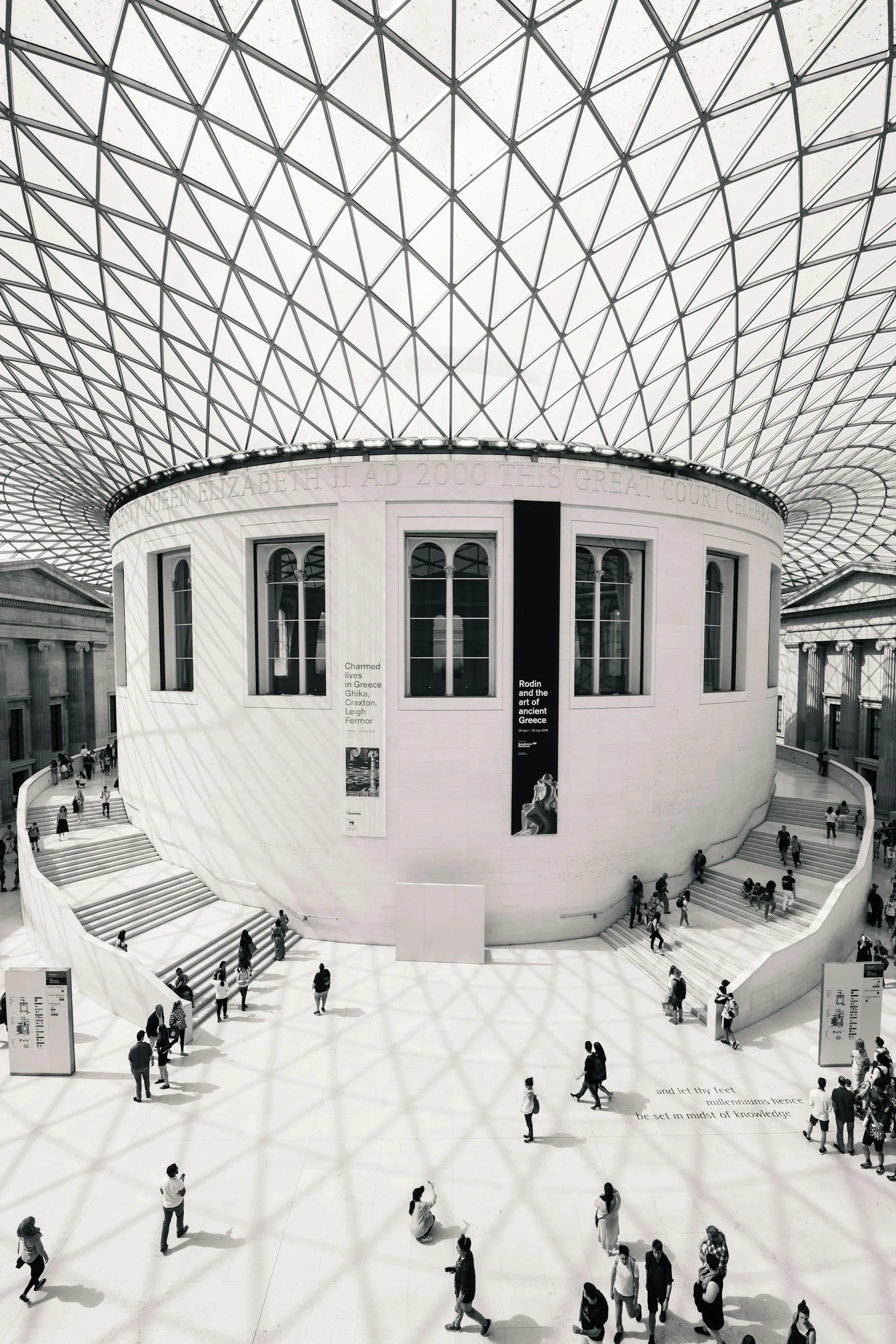 Interior view of a modern, spacious cultural or historical building with a large glass ceiling and a circular white wall. The space has multiple levels with stairs, and people are walking around, some taking photos and reading information displayed on boards.