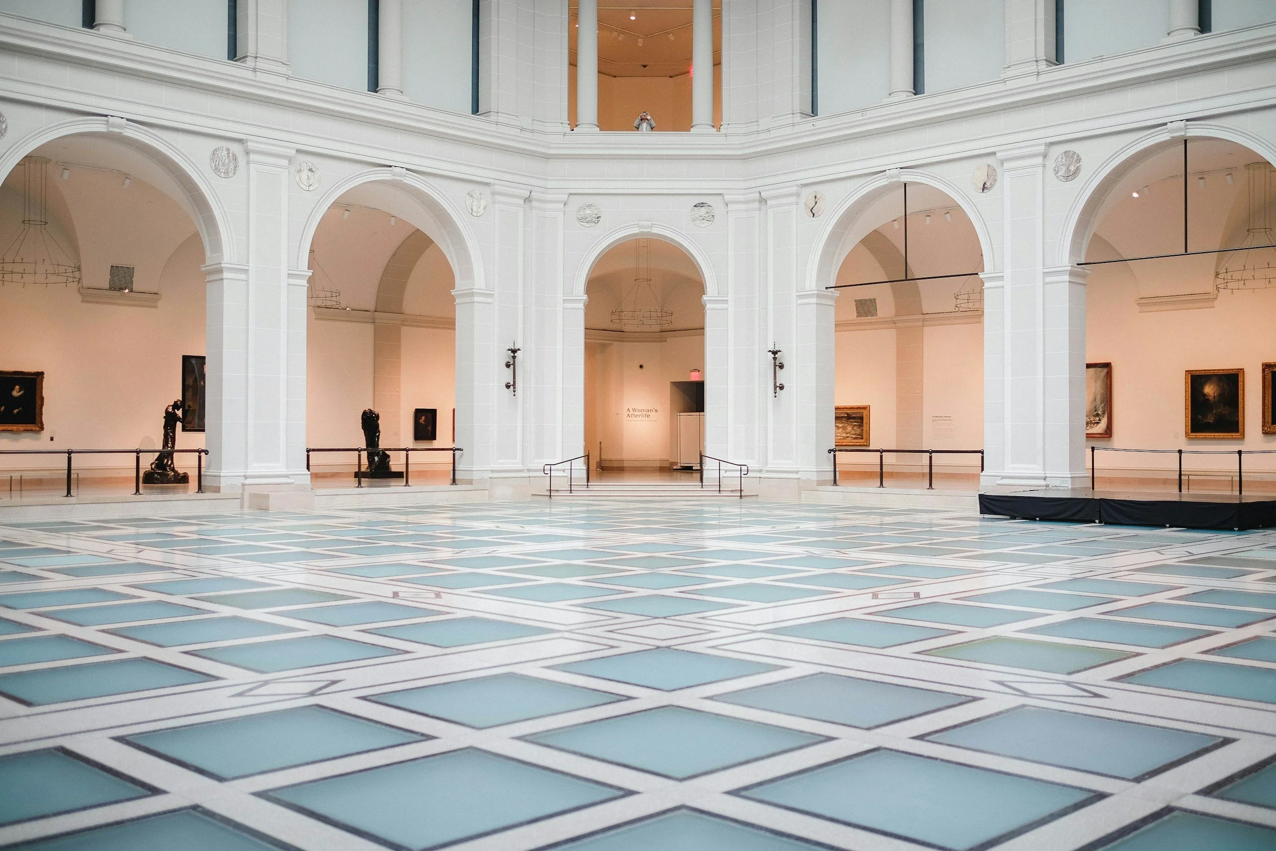 Interior of a museum with arched hallways, sculptures, and paintings displayed on the upper walls, with a person taking a photo from the balcony.