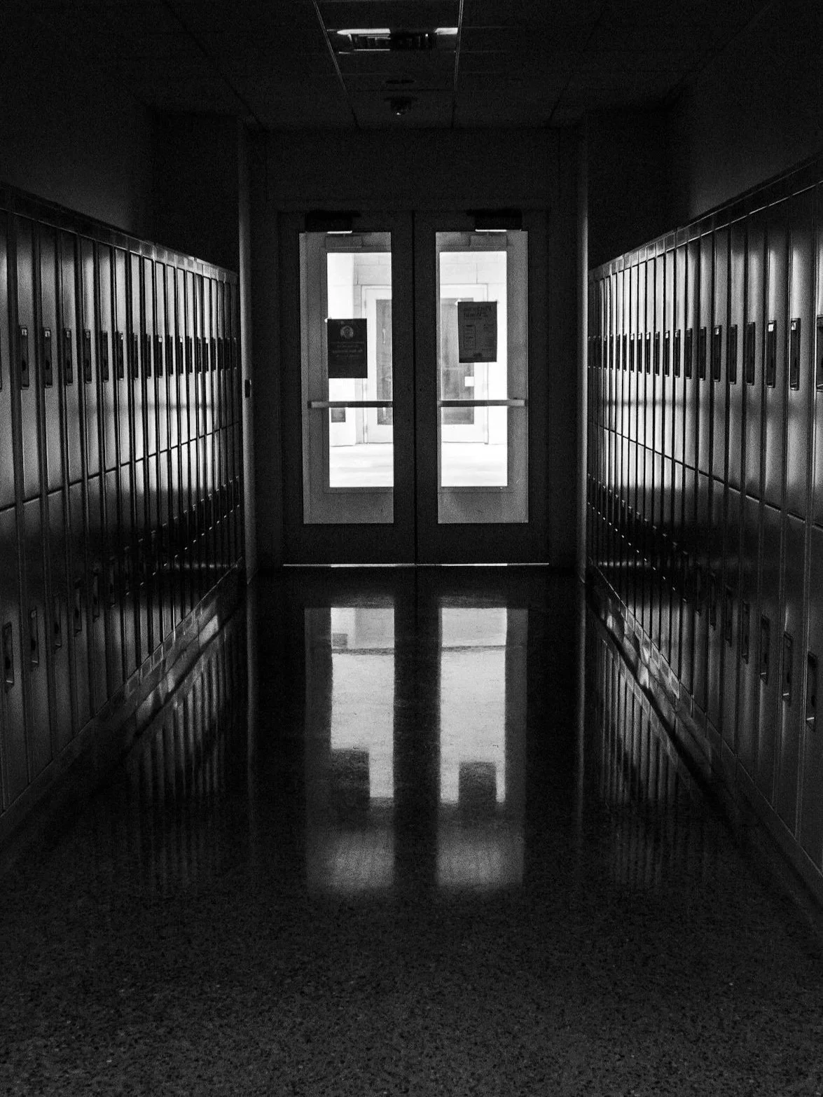 Black and white photo of school hallway with lockers on both sides and glass double doors at the end, with sunlight shining through.