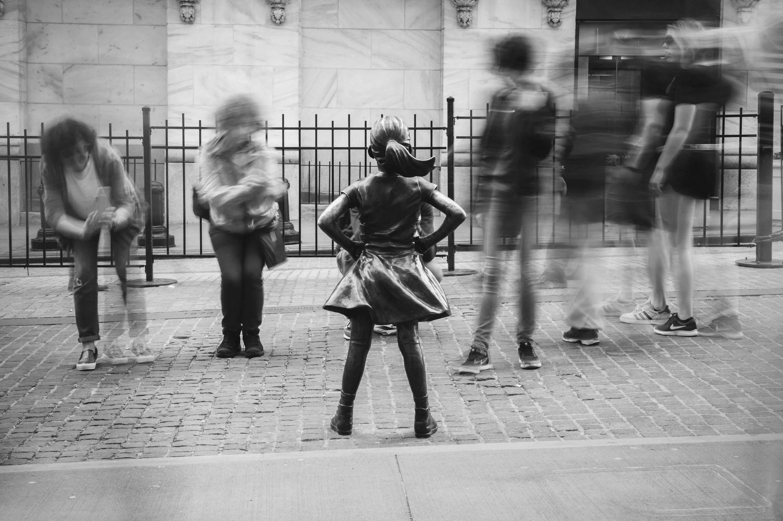 Black-and-white photo of children sitting on and standing around a statue of a girl in a dress, with blurred figures of people walking past in the background on a city sidewalk.