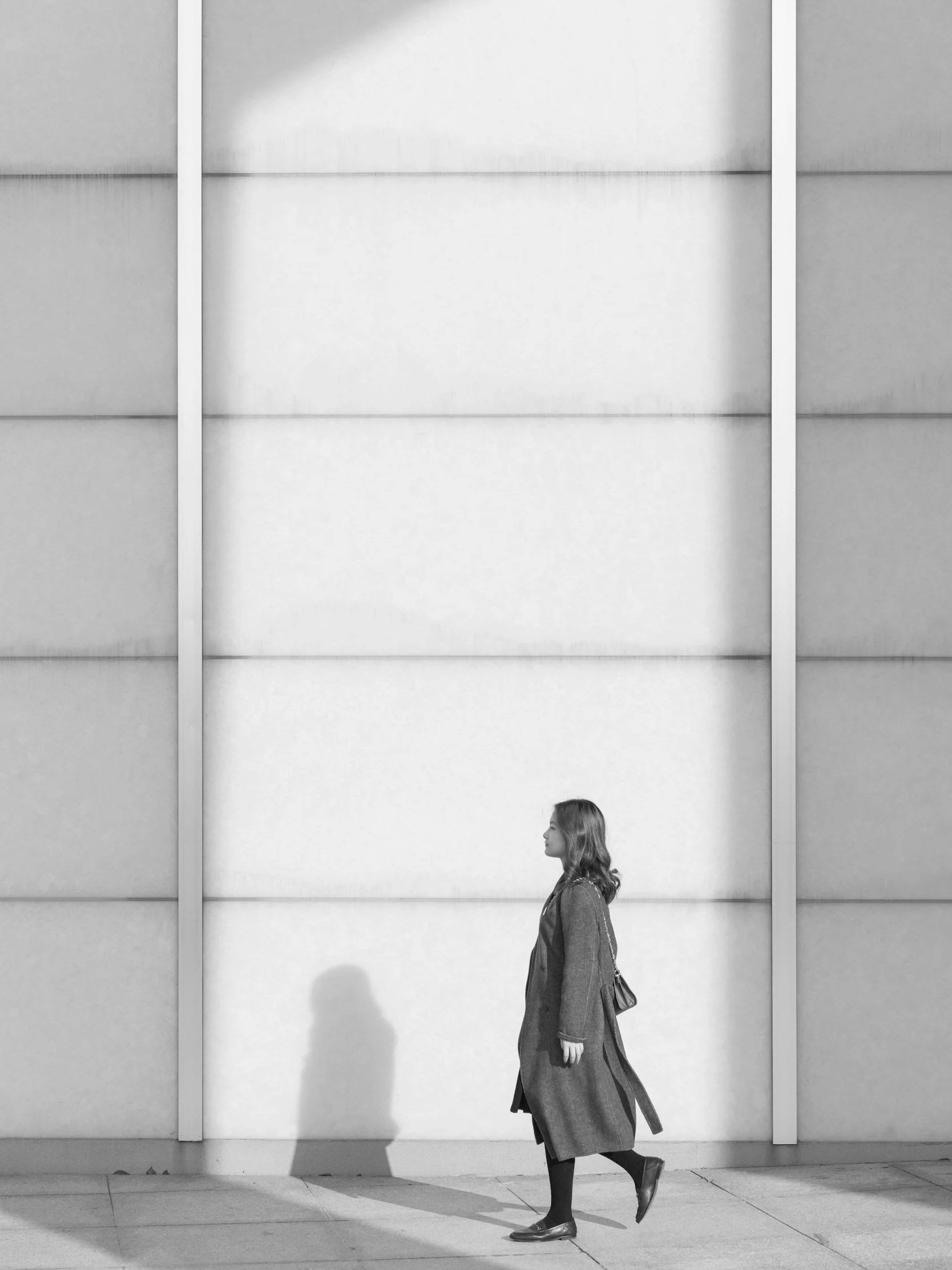 A woman walking on a sidewalk with sunlight casting a shadow on the wall behind her.