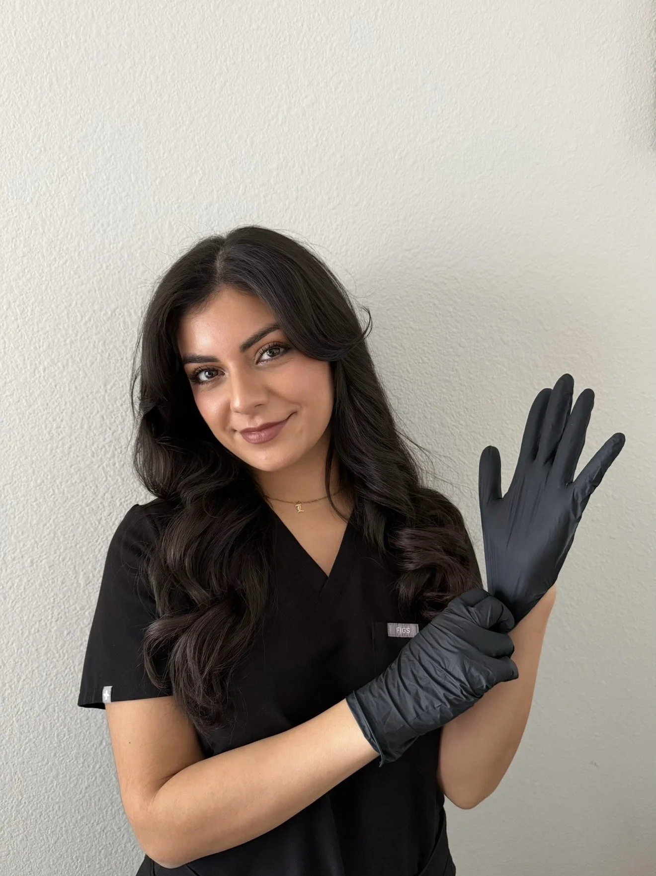 A young woman with long dark hair, wearing black scrubs and black gloves, smiling and waving with her left hand against a plain white wall.