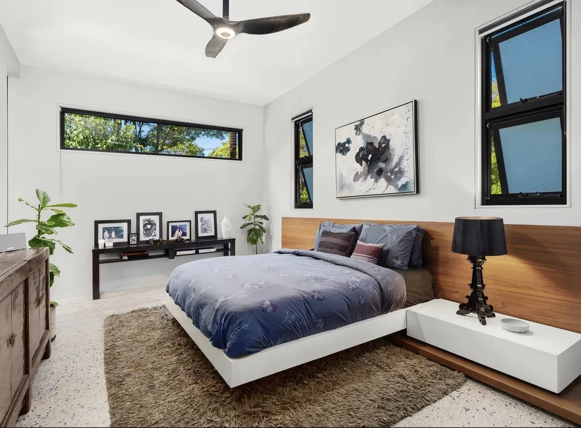 Modern bedroom with white walls, wooden accents, black-framed windows, a bed with gray and purple bedding, a black table lamp, framed photos and plants, and a ceiling fan.