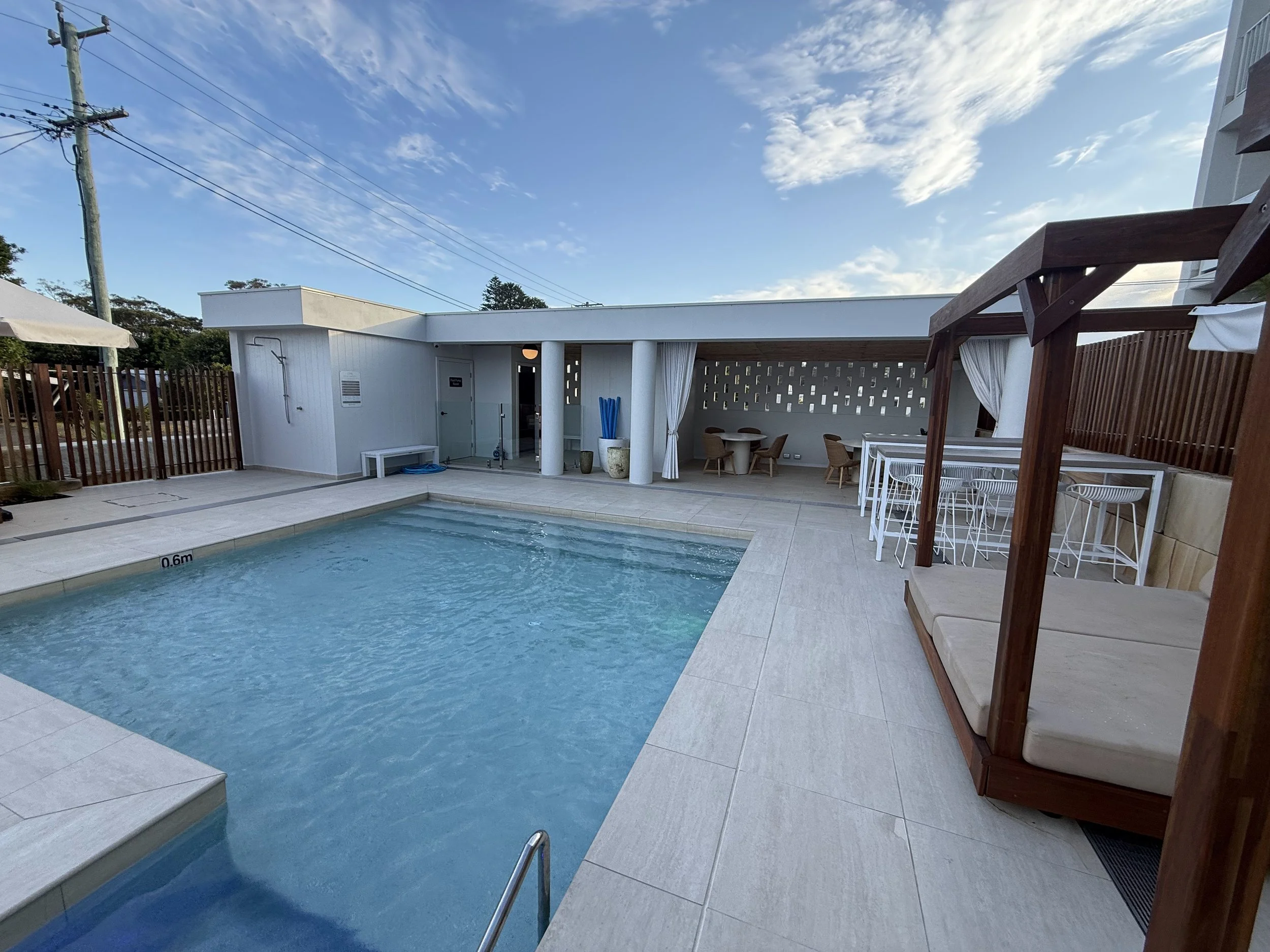 A swimming pool with a deck, outdoor furniture, a wooden shaded seating area with white curtains, and a modern building with a white wall and sliding doors, under a partly cloudy sky.