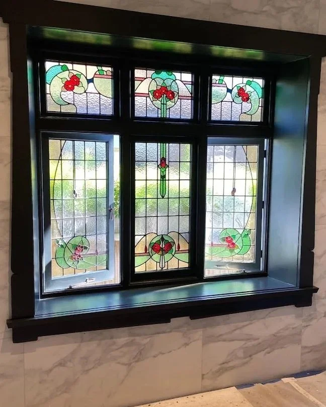 Colorful stained glass window with floral and vine motifs, set in a black frame on a stone wall.