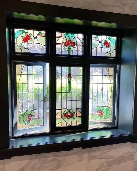 A decorative stained glass window with floral and vine patterns, featuring green and red colors, set in a dark frame.