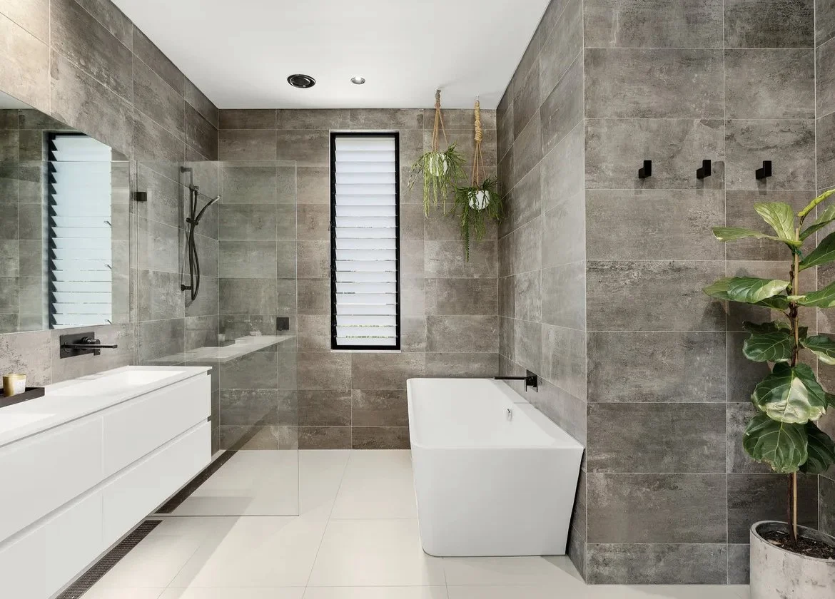Modern bathroom with gray tiled walls, a white bathtub, a floating white vanity with a sink, black fixtures, a glass shower enclosure, and indoor plants.