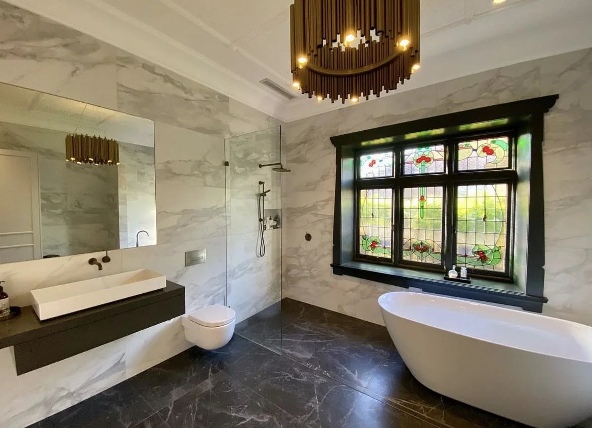 A modern bathroom with a large mirror, a white sink on a black floating vanity, a toilet, a glass-enclosed shower with a black showerhead, a window with stained glass design, a white freestanding bathtub, black trim around the window, a dark marble floor, and a black ceiling light fixture.