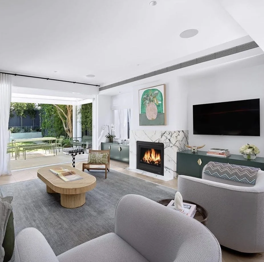 Modern living room with a fireplace, wall-mounted TV, gray sofa, armchair, coffee table, and an open sliding door leading to a patio with outdoor furniture and greenery.