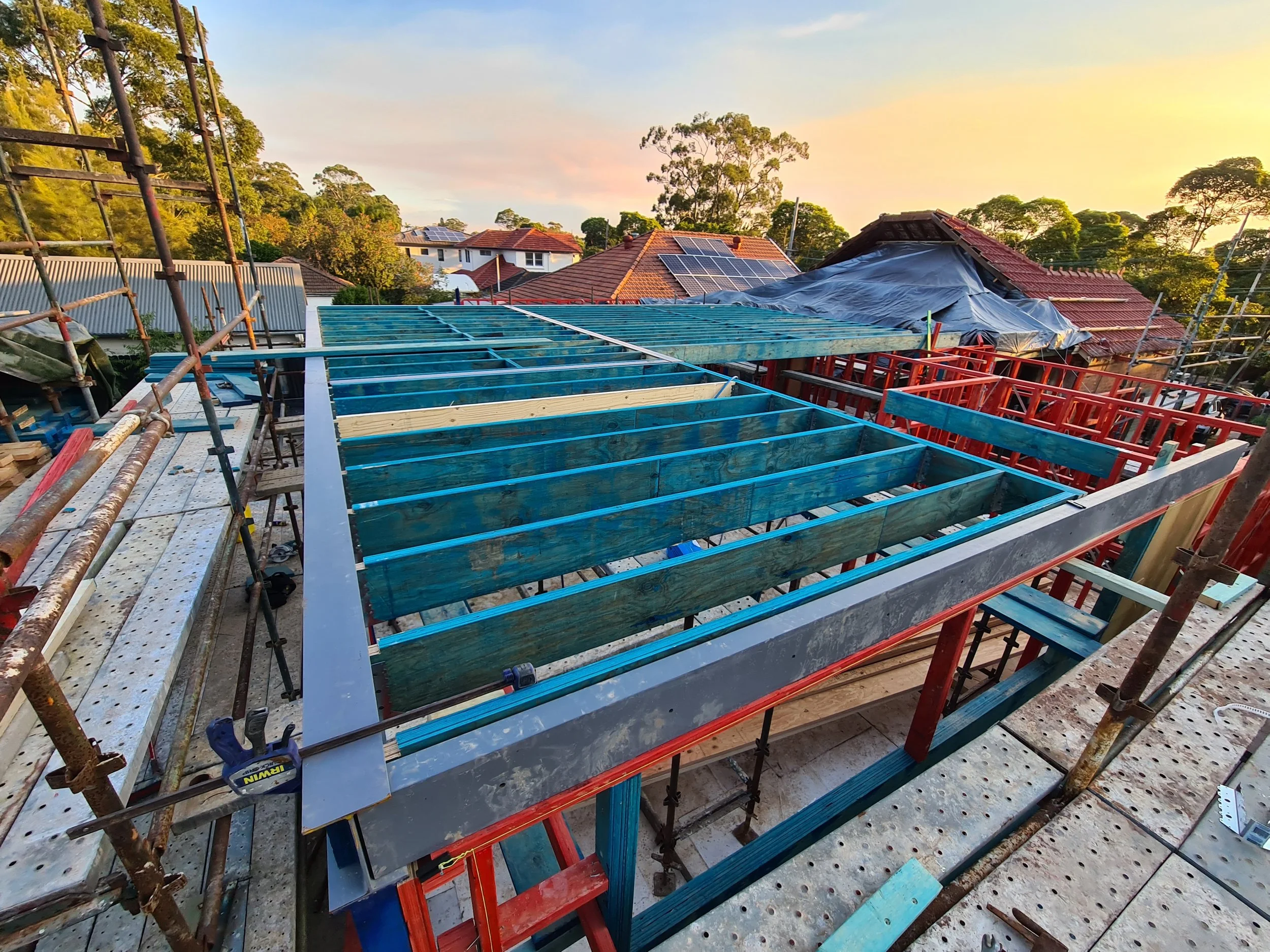 Construction site on a residential roof with scaffolding, blue framing for a new roof section, solar panels installed on neighboring roofs, and a sunset sky.