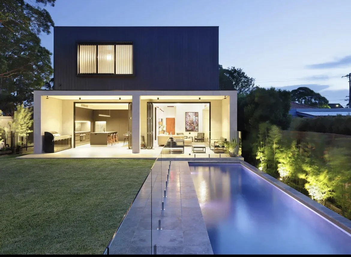 Modern two-story house with an illuminated outdoor living space, swimming pool, and landscaped yard at dusk.