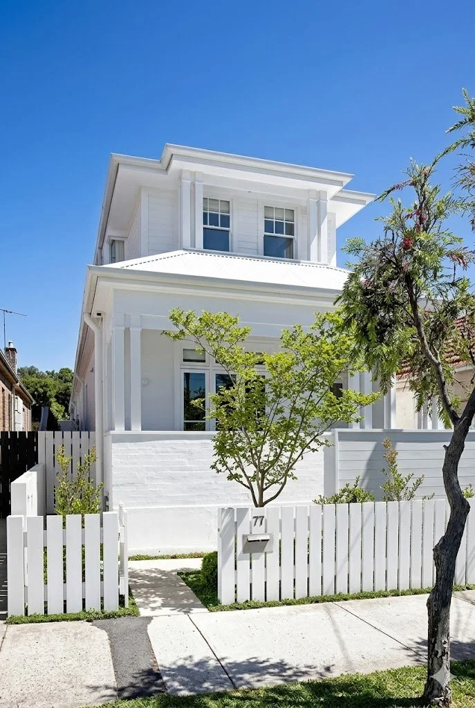 A white two-story house with a front porch, surrounded by a small white picket fence, with trees and a blue sky in the background.