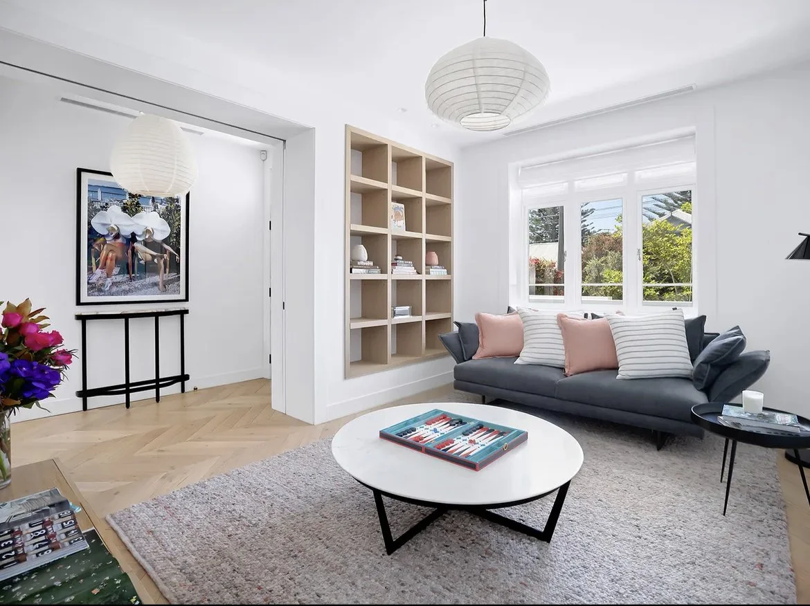 Bright living room with a gray sofa adorned with striped and pink pillows, a white round coffee table with a board game, a black side table with a candle, a large window with white blinds, a white ceiling with hanging paper lantern lights, a light-colored wooden bookshelf filled with books, and a colorful bouquet of flowers on a side table.