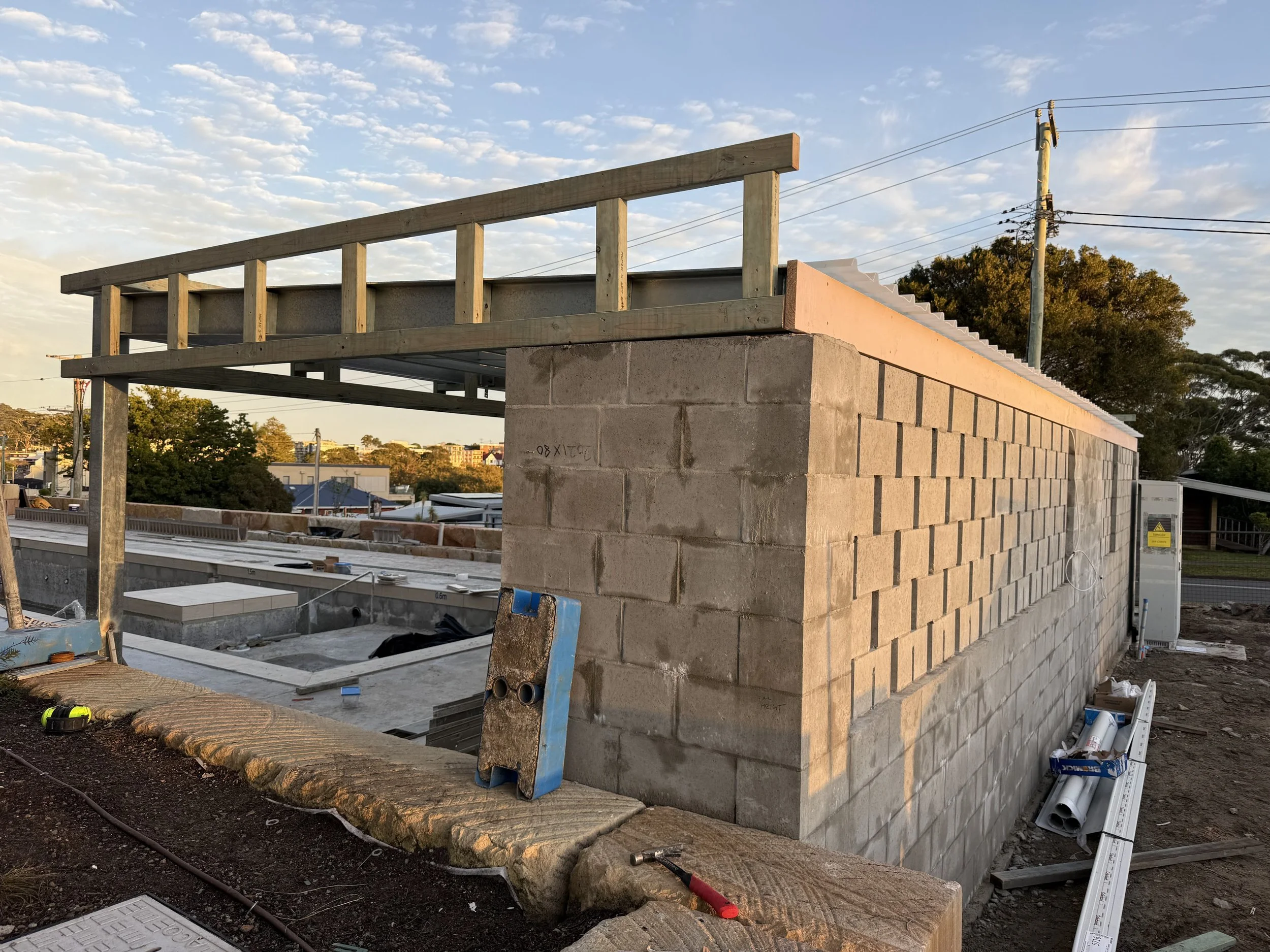 Construction site with a brick wall under construction, wooden framework on top, and construction tools and materials nearby, during a clear day with scattered clouds.