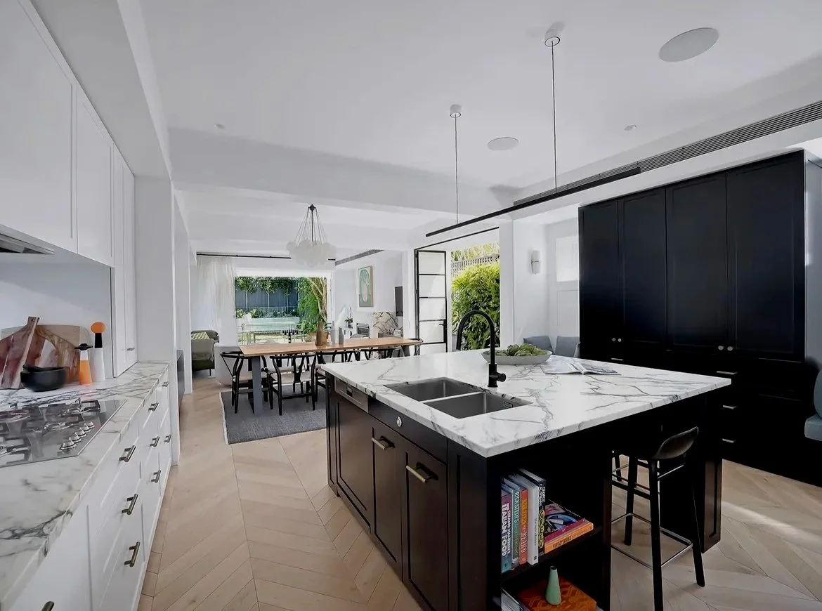 Modern open-plan kitchen with black and white cabinetry, a marble island with a sink, and a view of a dining area and living room with natural light and greenery outside.