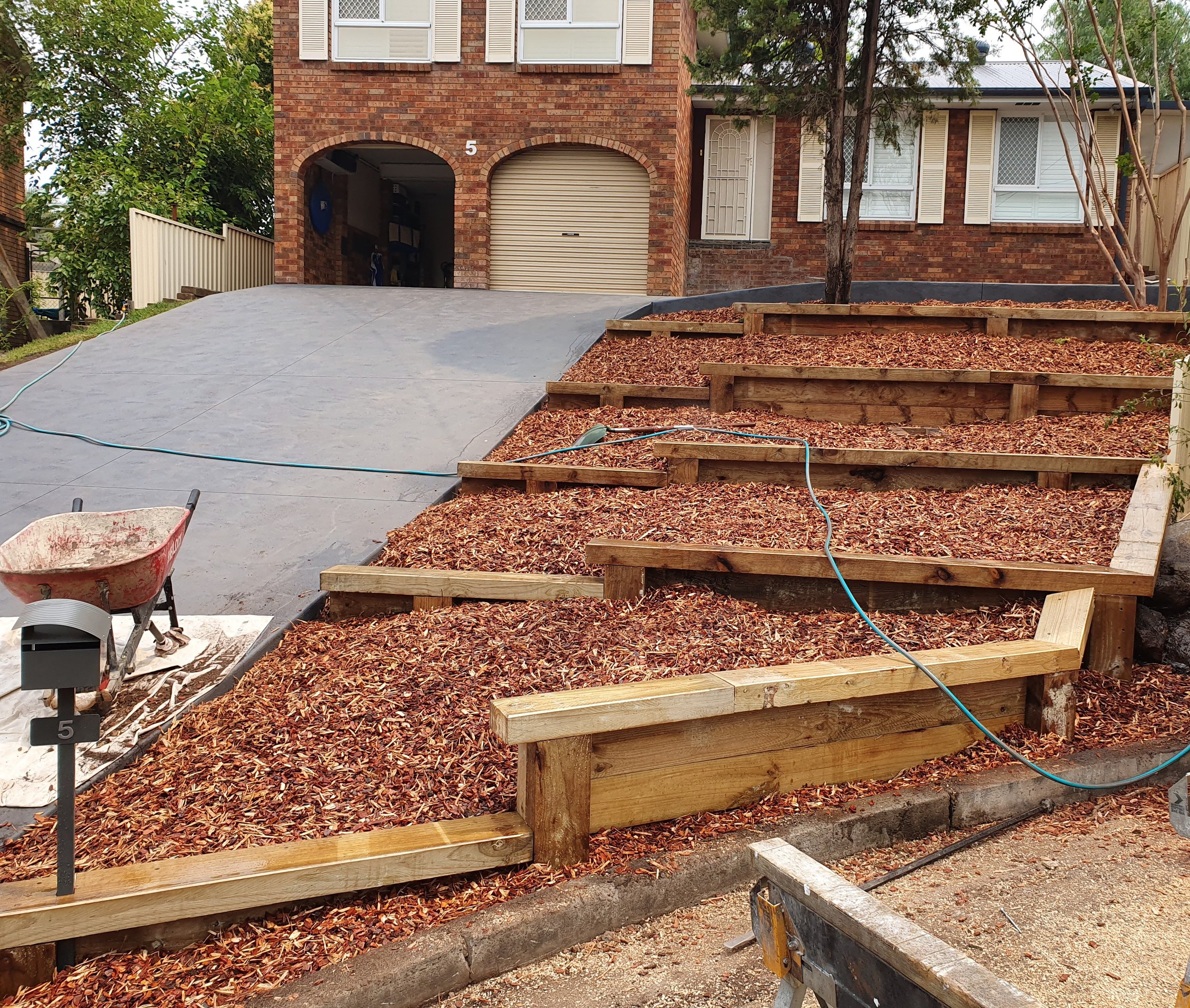 Residential backyard in the process of construction with a newly built wooden staircase and a sloped concrete driveway in front of a brick house. There are trees and leaf mulch surrounding the area.