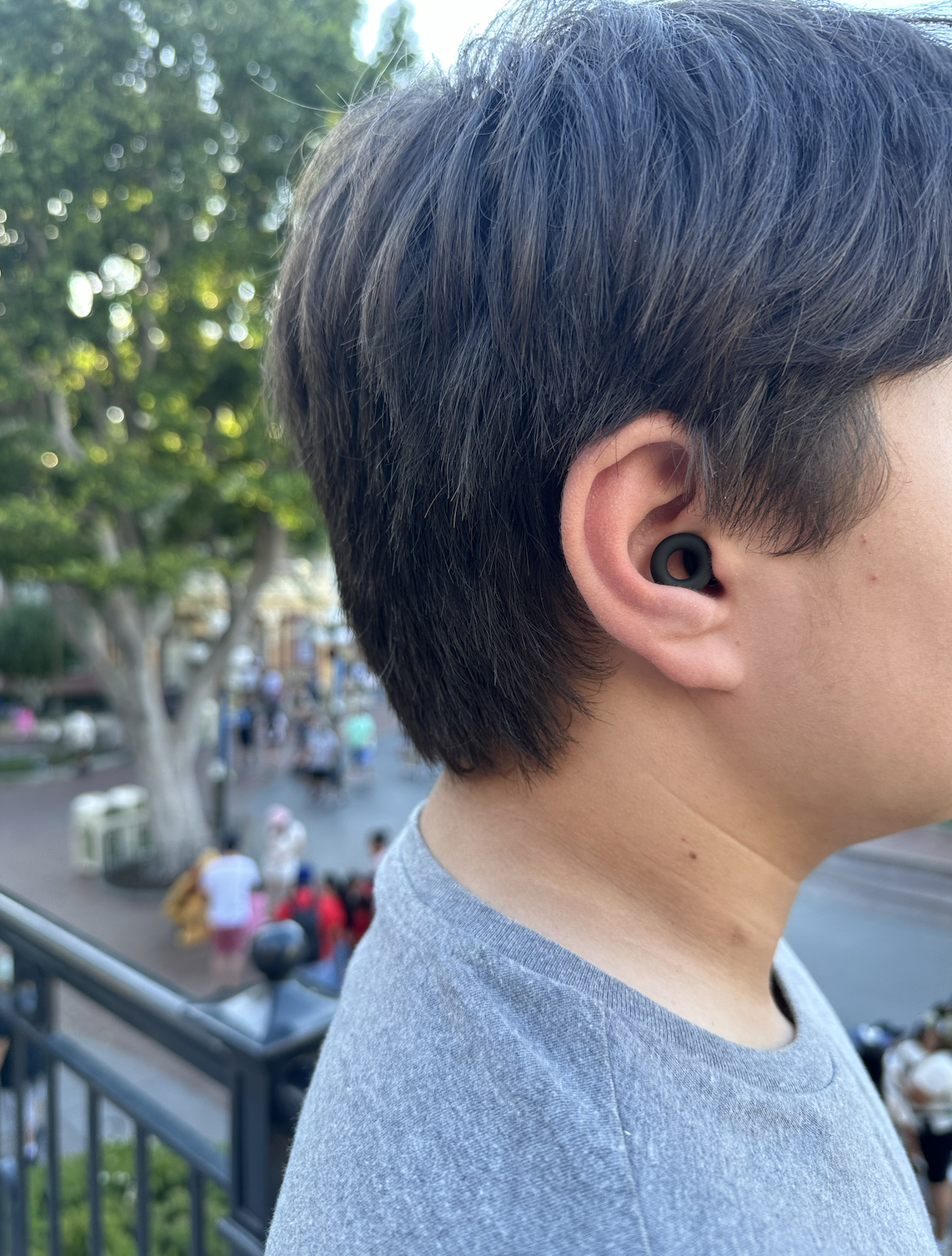 A person with short dark hair wearing a black wireless earbud, standing outdoors with a blurred background of trees, people, and a street scene.