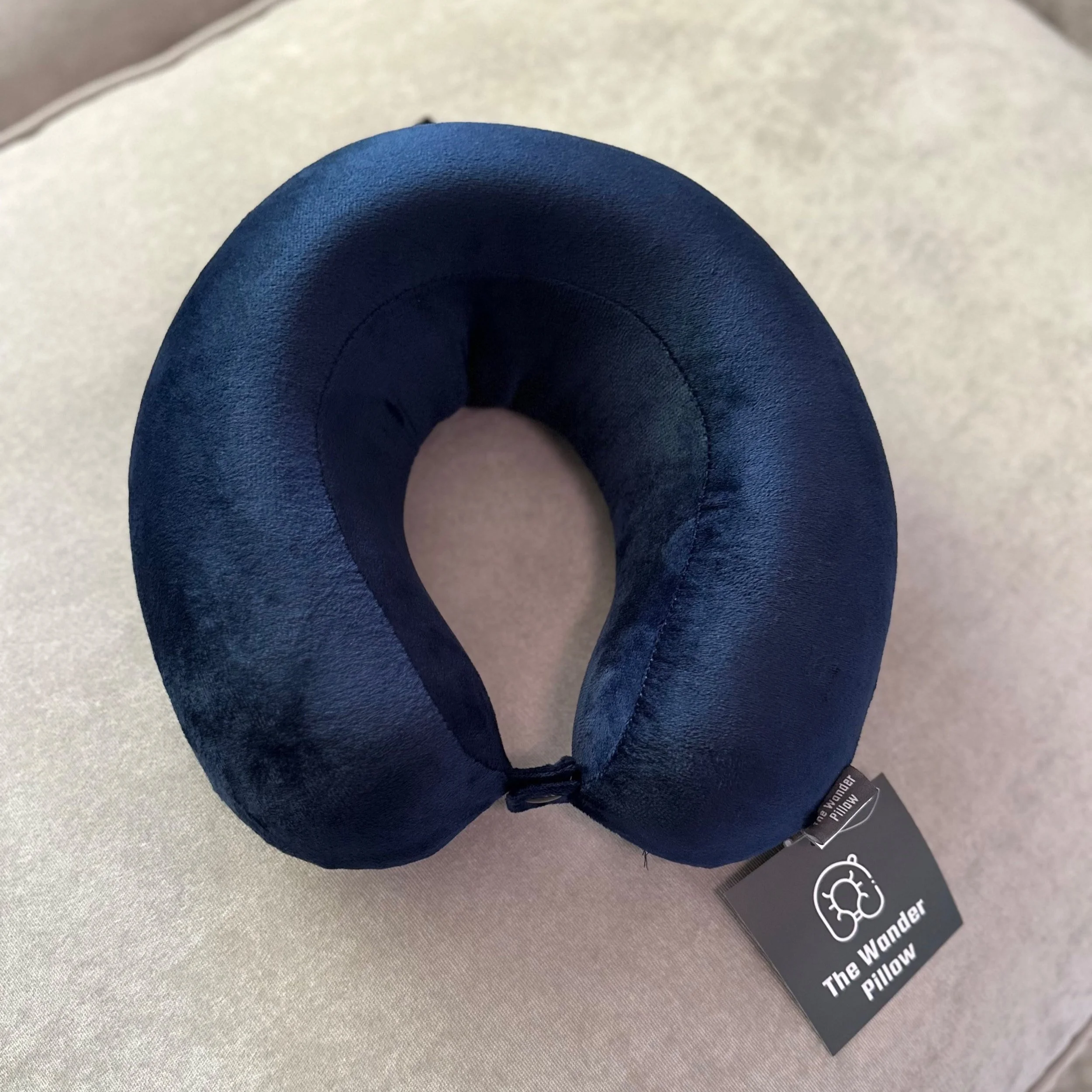 A dark blue U-shaped neck pillow resting on a light gray fabric surface, with a black tag displaying the brand name and a warning label partially visible.
