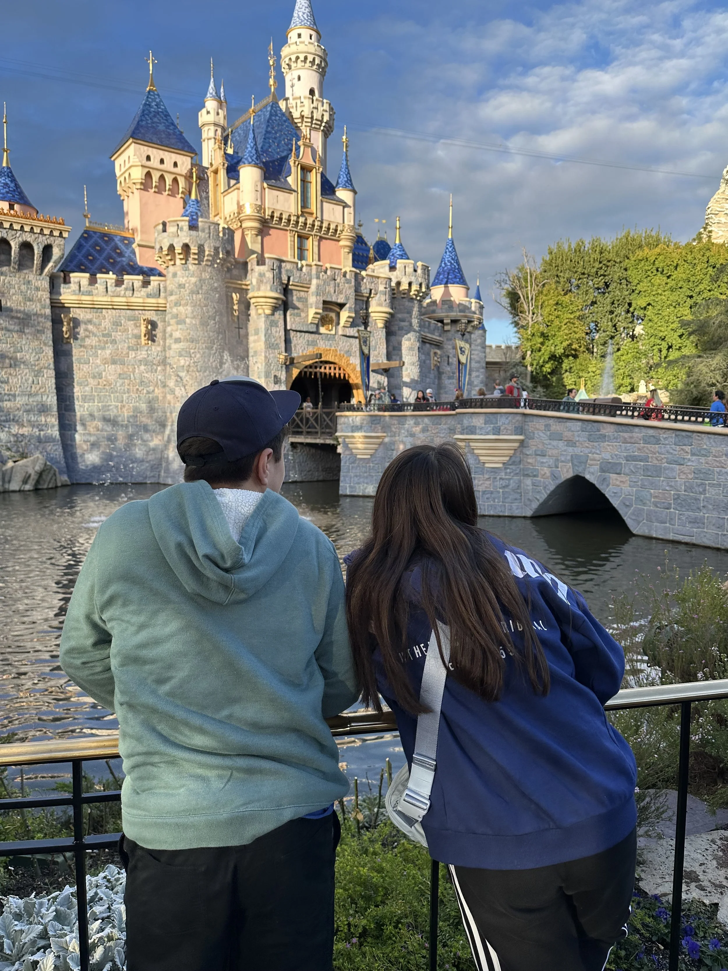 Two people stand by a railing looking at Sleeping Beauty Castle at Disneyland, with a moat in the foreground, lush green trees to the right, and a partly cloudy sky in the background.